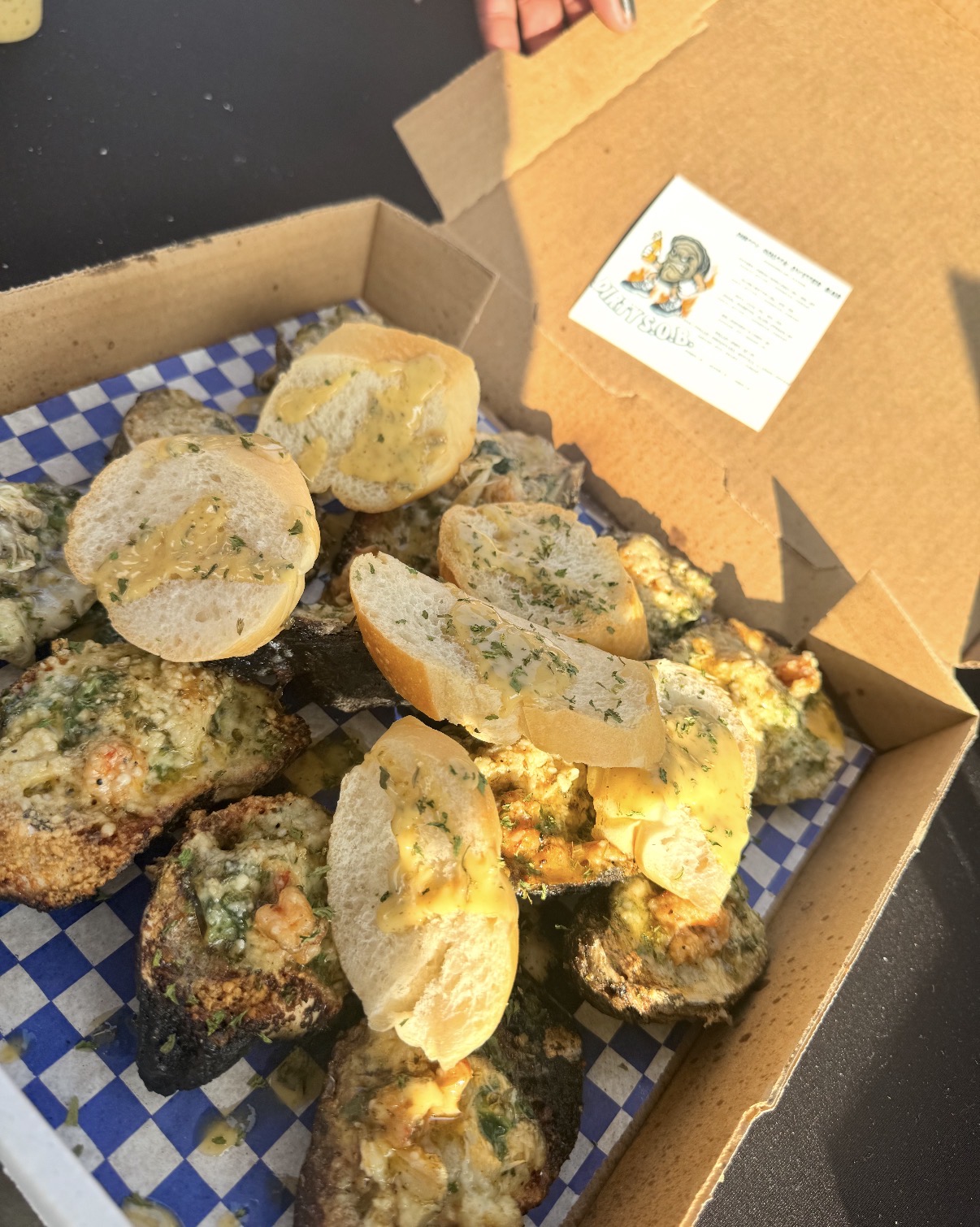 Open cardboard box containing fried food items with a yellowish coating, on blue and white checkered paper, with a small card inside.