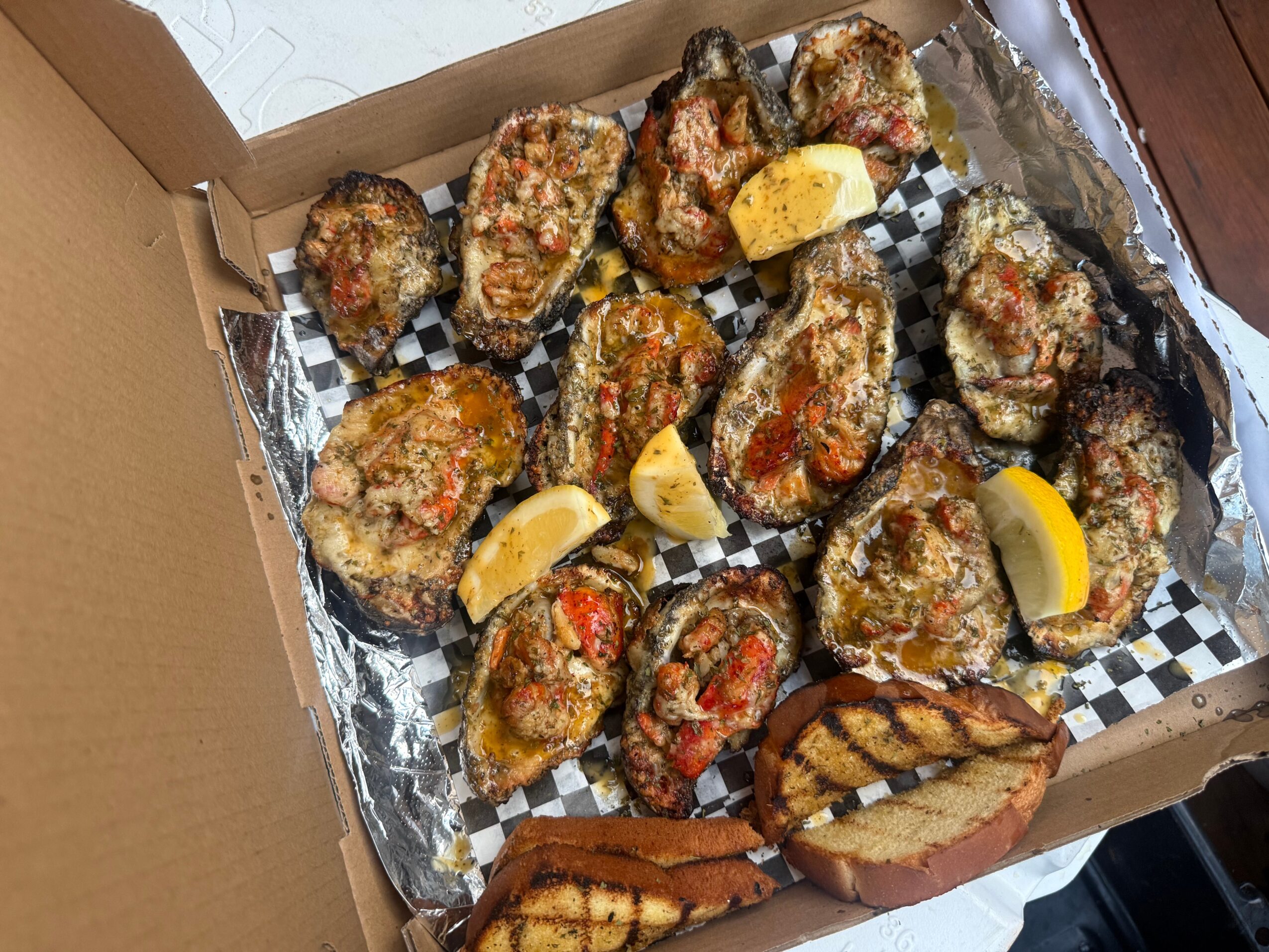 Open pizza box with fried oysters and lemon wedges, lined with foil and checkered paper, on a wooden surface.