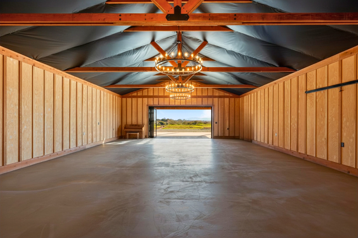 Empty room with wooden walls, a chandelier, and a view outside through an open door, concrete floor, ceiling with exposed beams.