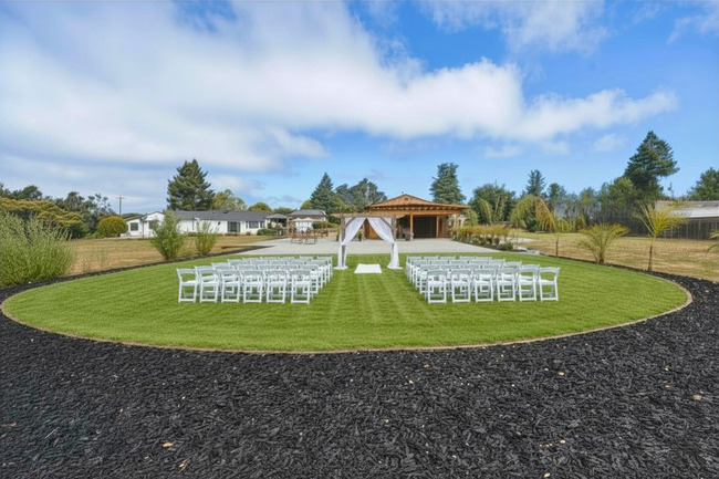 Outdoor event space with white chairs arranged in rows on a circular grassy area, a wooden pavilion, and a blue sky with clouds.