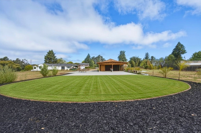 A circular grassy area surrounded by black gravel, with a wooden pavilion and trees in the background under a partly cloudy sky.