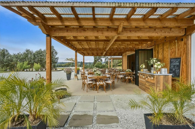 Outdoor patio with wooden roof, tables, chairs, and plants, overlooking a body of water and trees in the background.