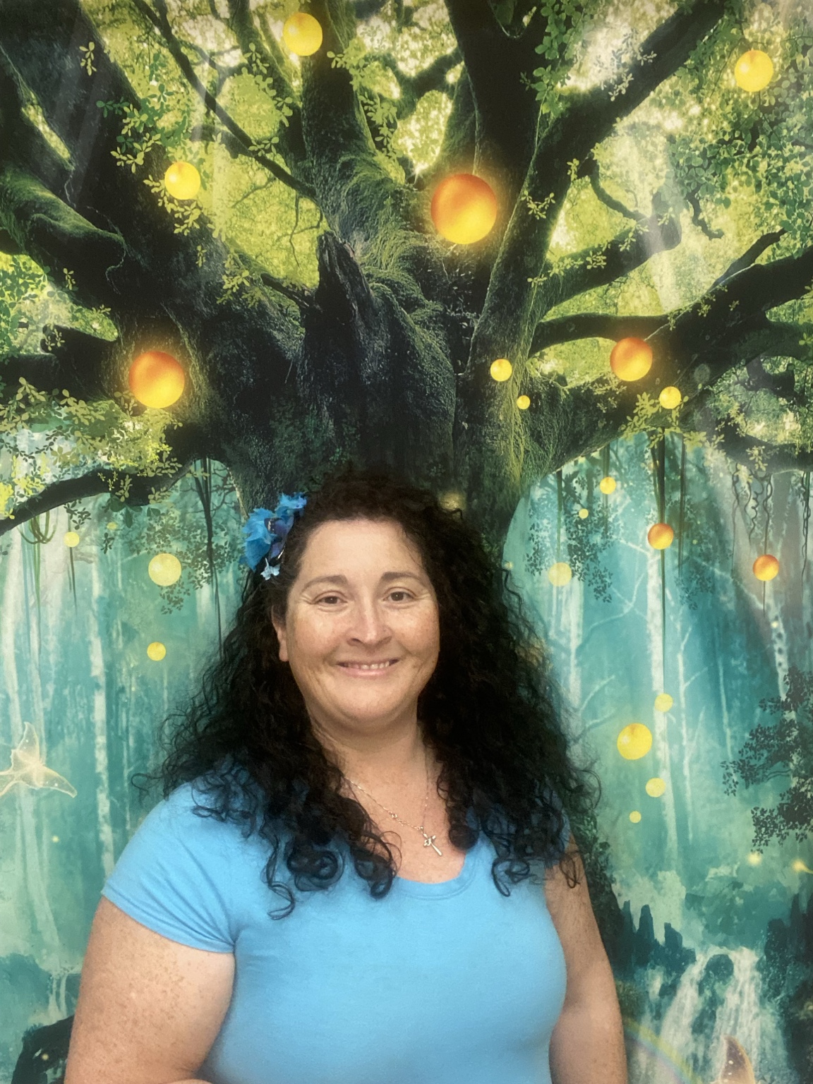 Woman with curly dark hair smiling in front of a colorful forest background with glowing orbs.
