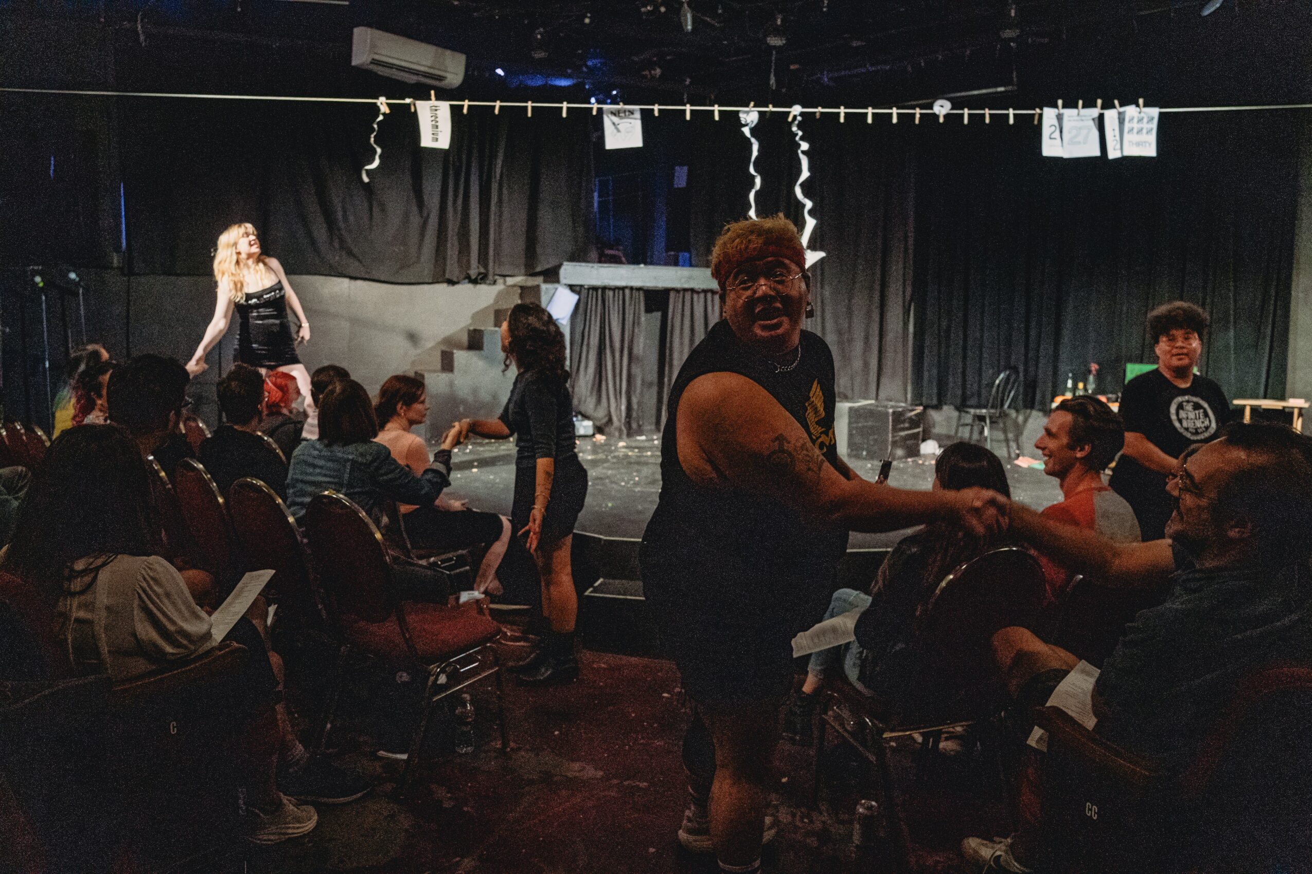 Person standing and shaking hands with another person in a room with seated audience and stage background.