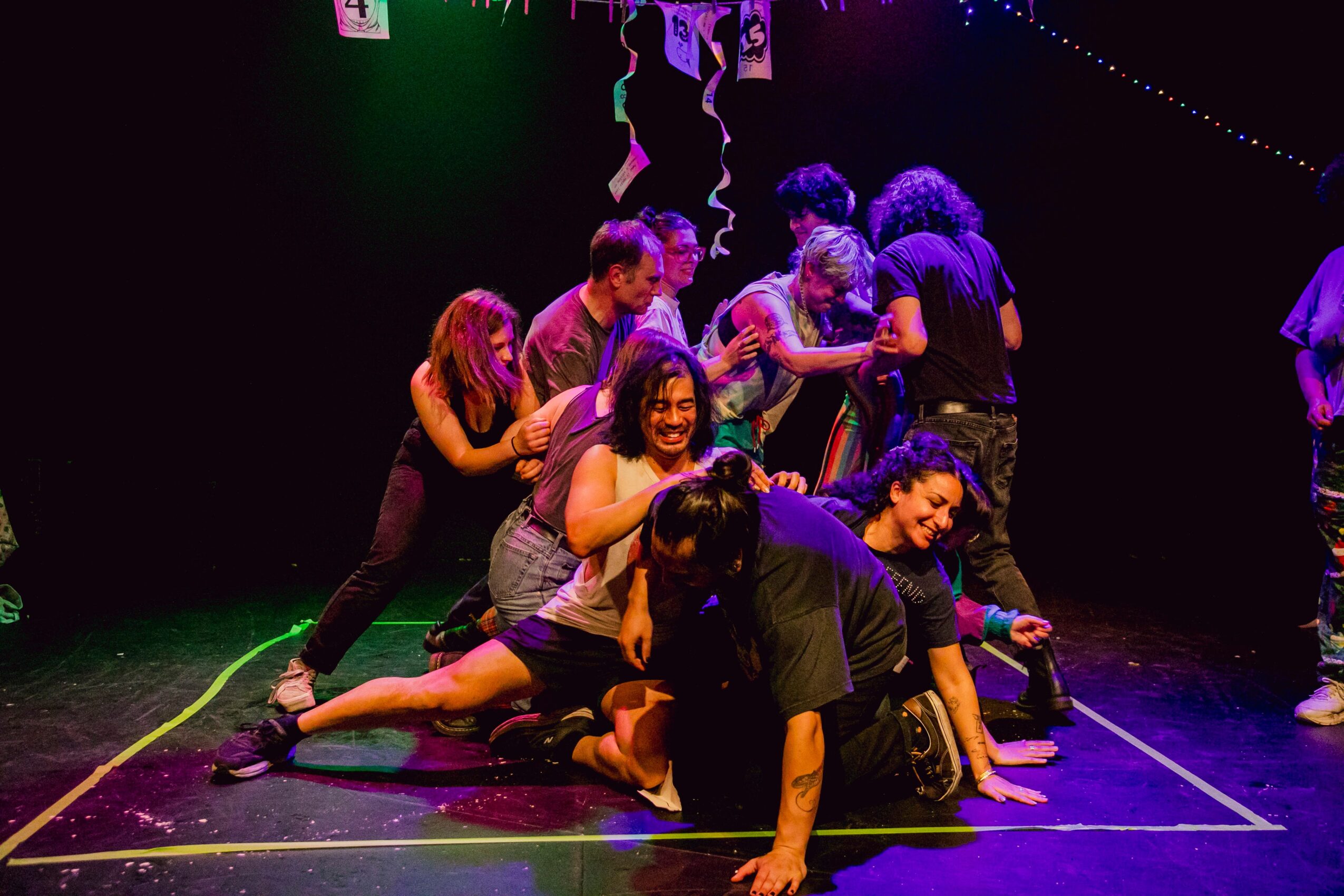 Group of people performing a dance or theatrical scene on stage with colorful lighting, some kneeling and others standing.