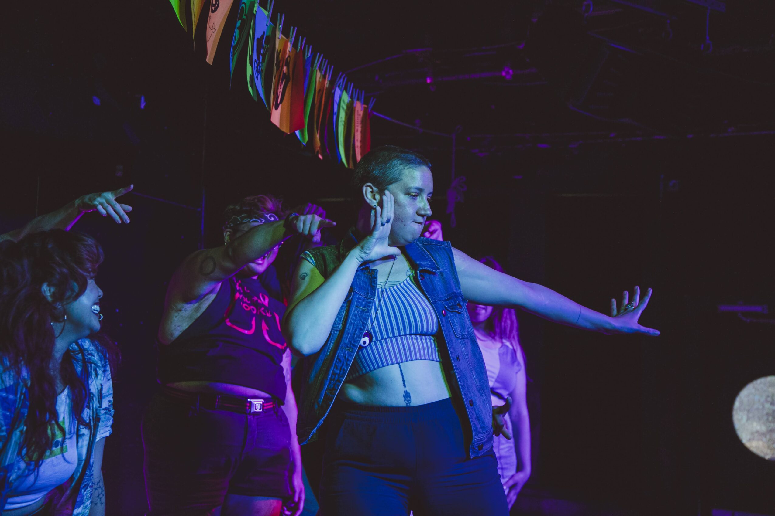 Group of people dancing in a dark, colorful setting with a woman in the center wearing a crop top and vest.