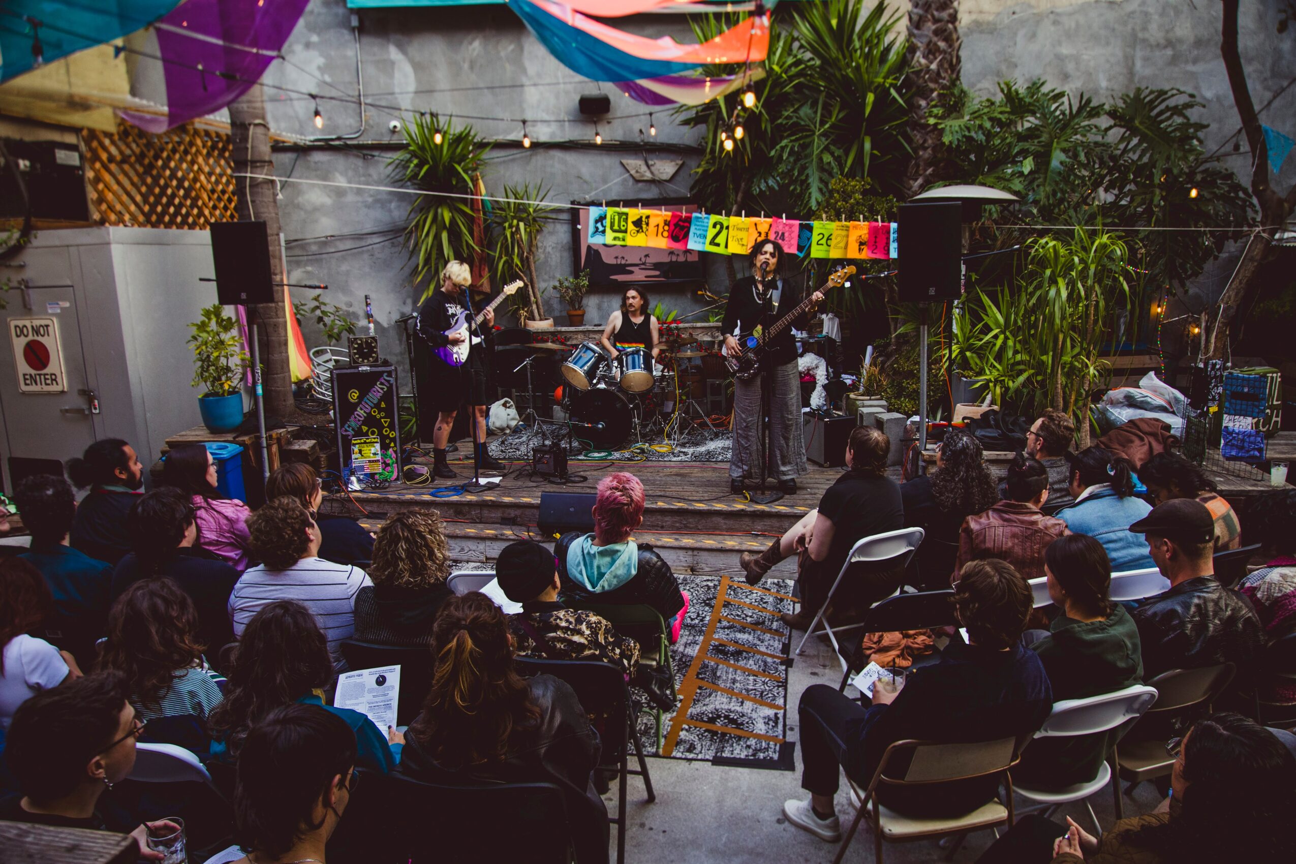 Outdoor concert with a band performing on stage, audience seated, colorful decorations, and lush greenery in the background.