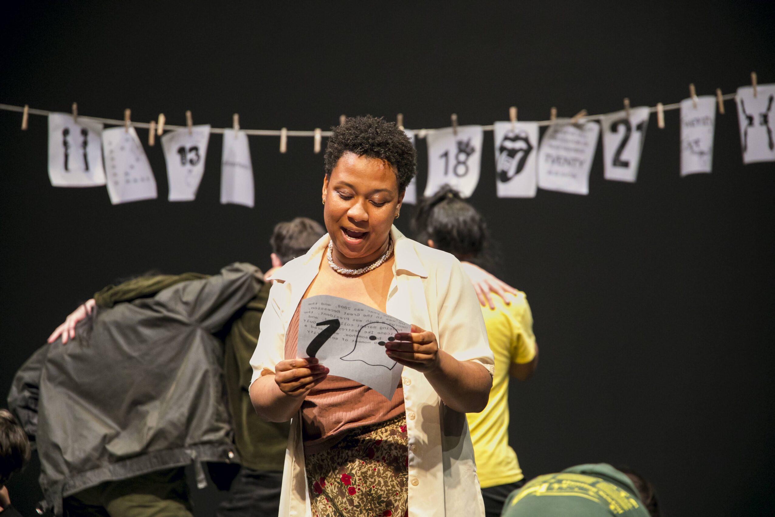 A woman reads from a paper on stage, with others in the background and papers hanging above. She appears engaged.