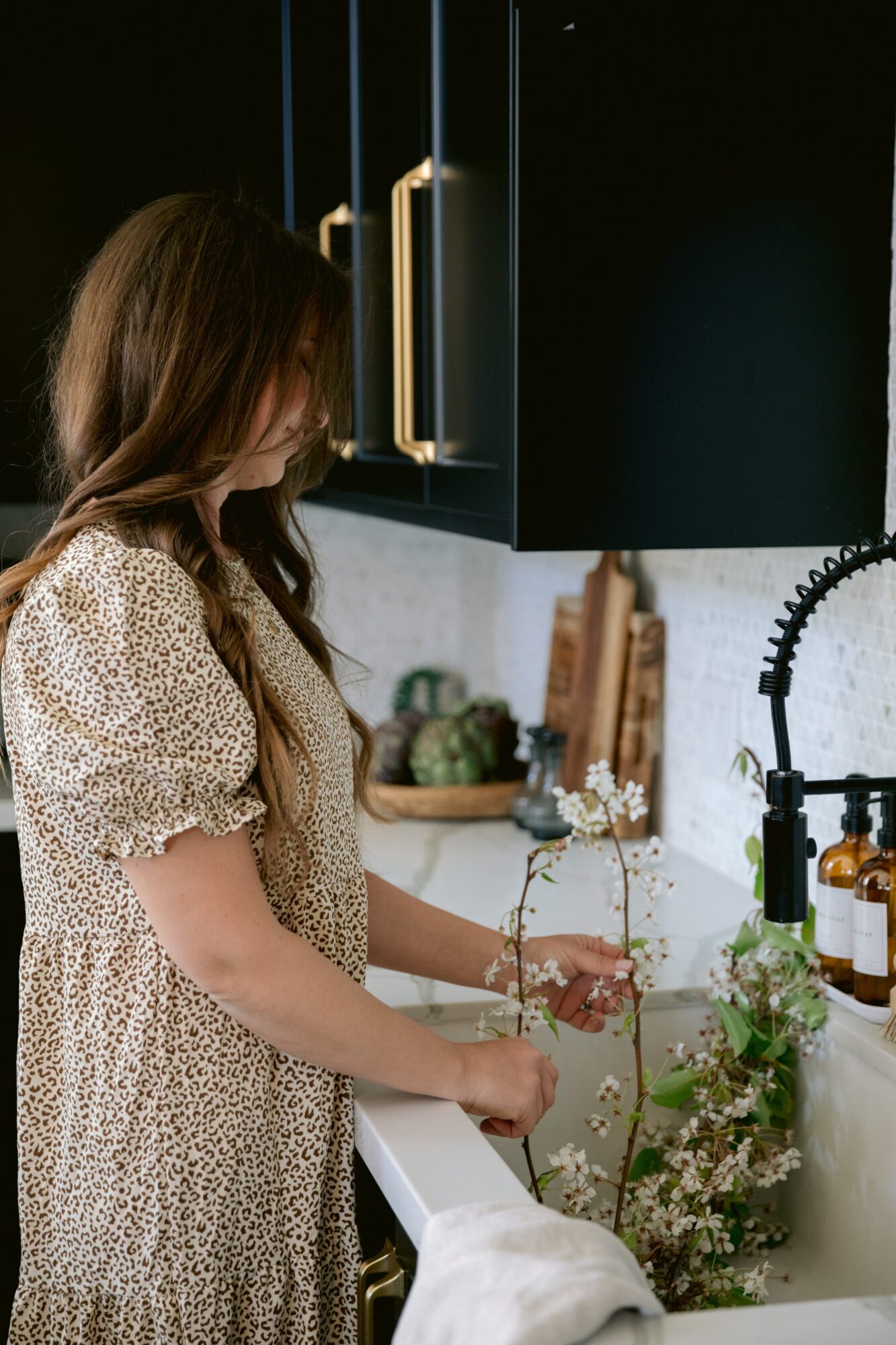Woman with long hair in a patterned dress arranging flowers in a kitchen sink, dark cabinets above, various items on counter.