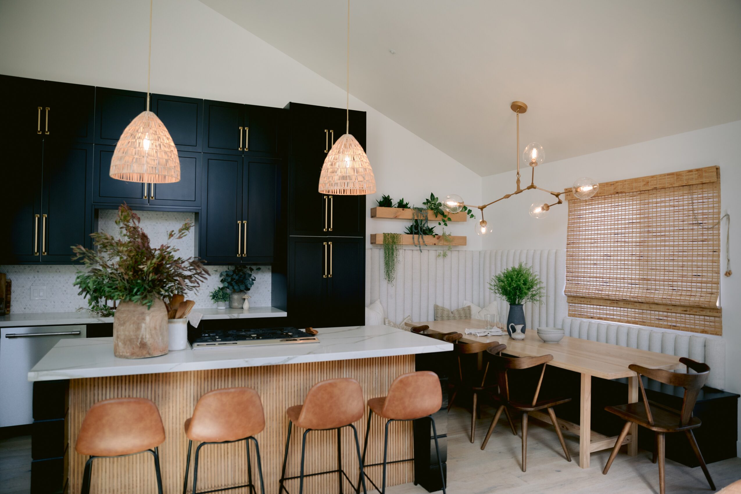 Kitchen with black cabinets, white countertop, hanging pendant lights, and a dining area with chairs and a window with woven shade.