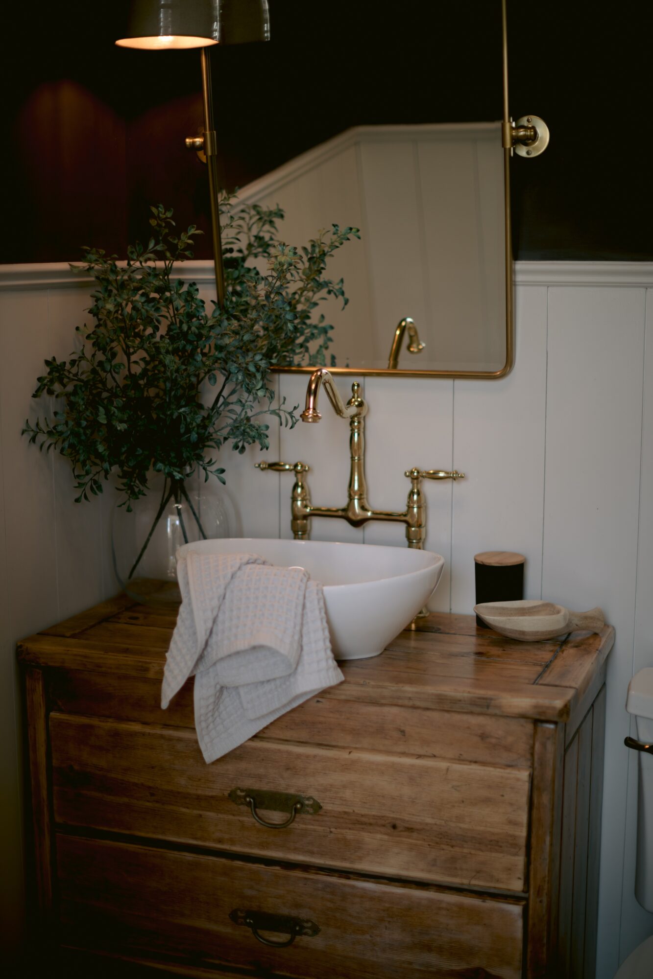 Bathroom sink with a mirror, plant, and gold fixtures on a wooden cabinet, with a towel and soap nearby.