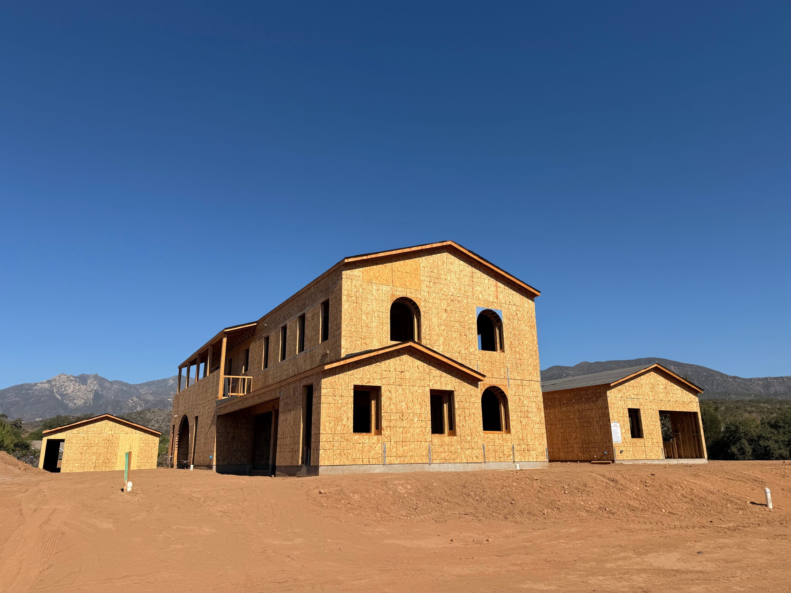 A large brick building with multiple windows and arches, surrounded by a dirt area and smaller structures, under a clear blue sky.