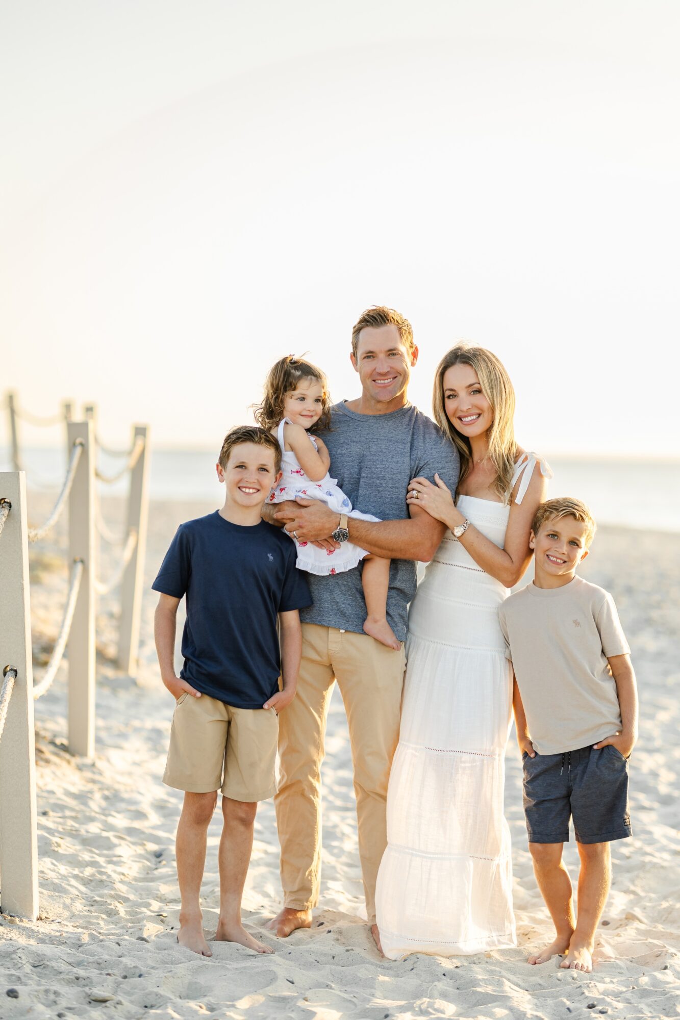 Family of five standing on beach, smiling, with ocean and sky in background.
