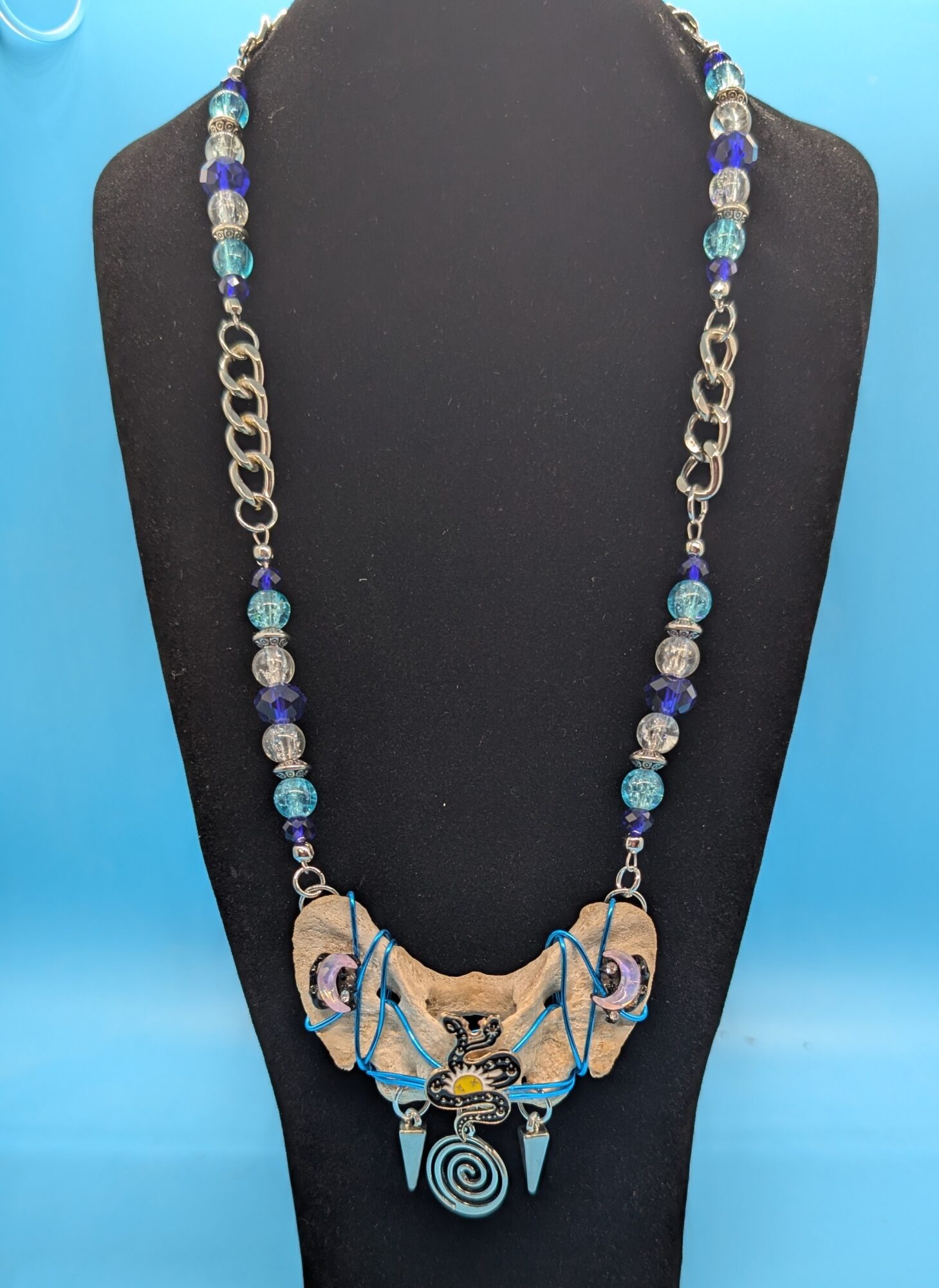 Beaded necklace with a large, ornate central pendant featuring a spiral design, on a black display bust.