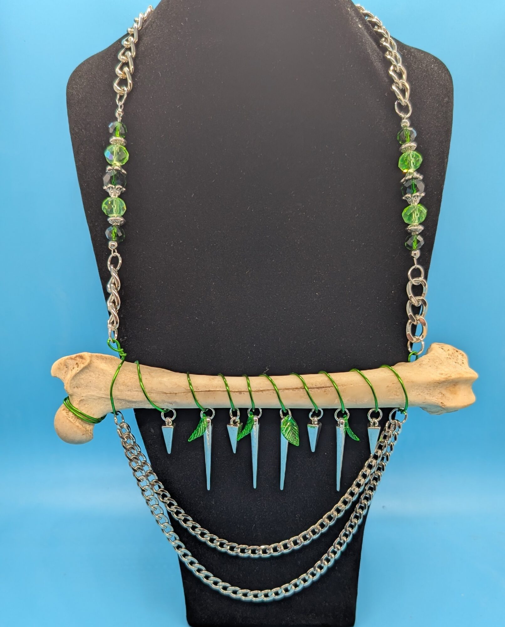 Necklace with green beads, silver chain, and hanging blue and green spikes displayed on a black bust against a blue background.
