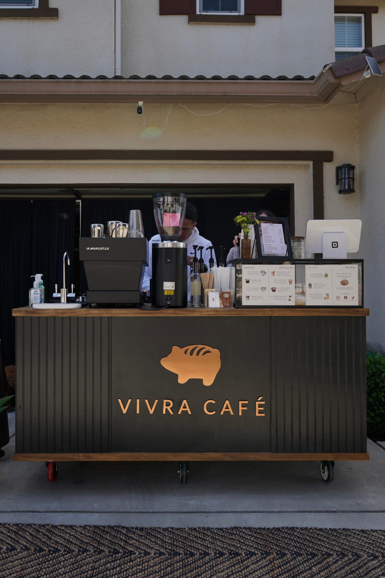 Outdoor coffee stand with blender, cups, and boxes in front of a house, Vivra Café logo with fish icon, and a person behind.