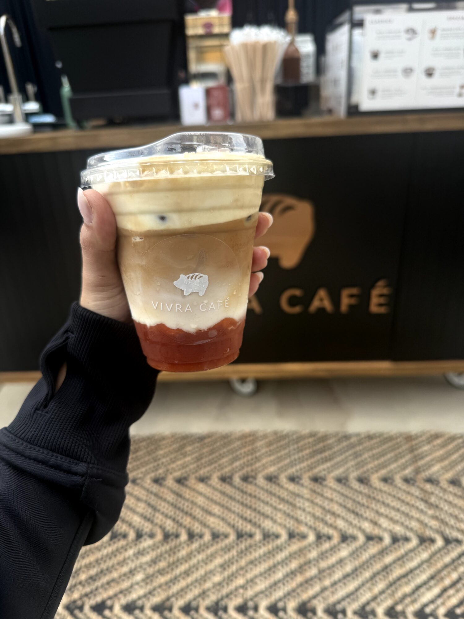 Hand holding layered coffee drink in clear cup with a plastic lid, background of a cafe counter and sign.
