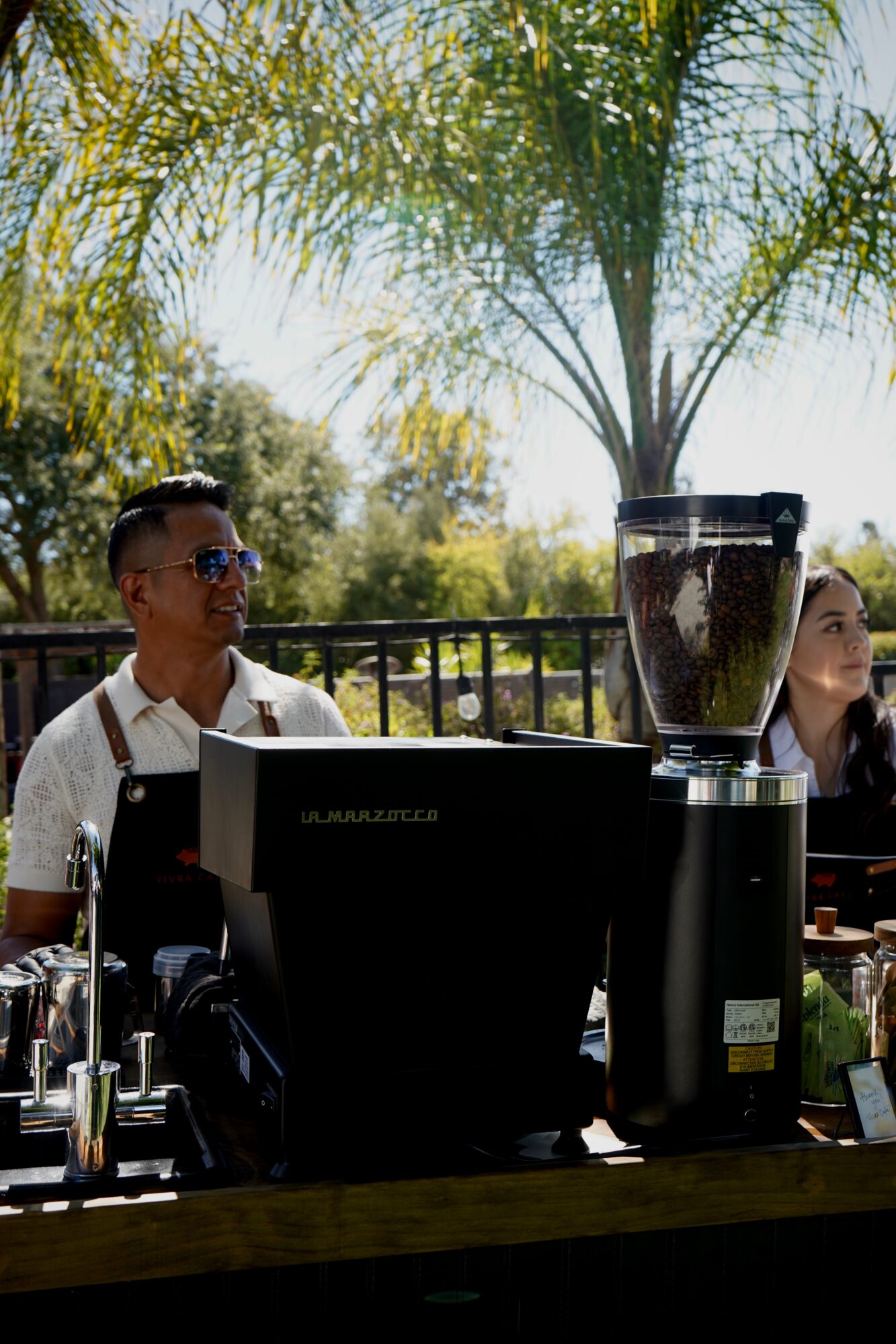 Two people behind a counter outdoors with a blender, trees, and sunlight in the background.