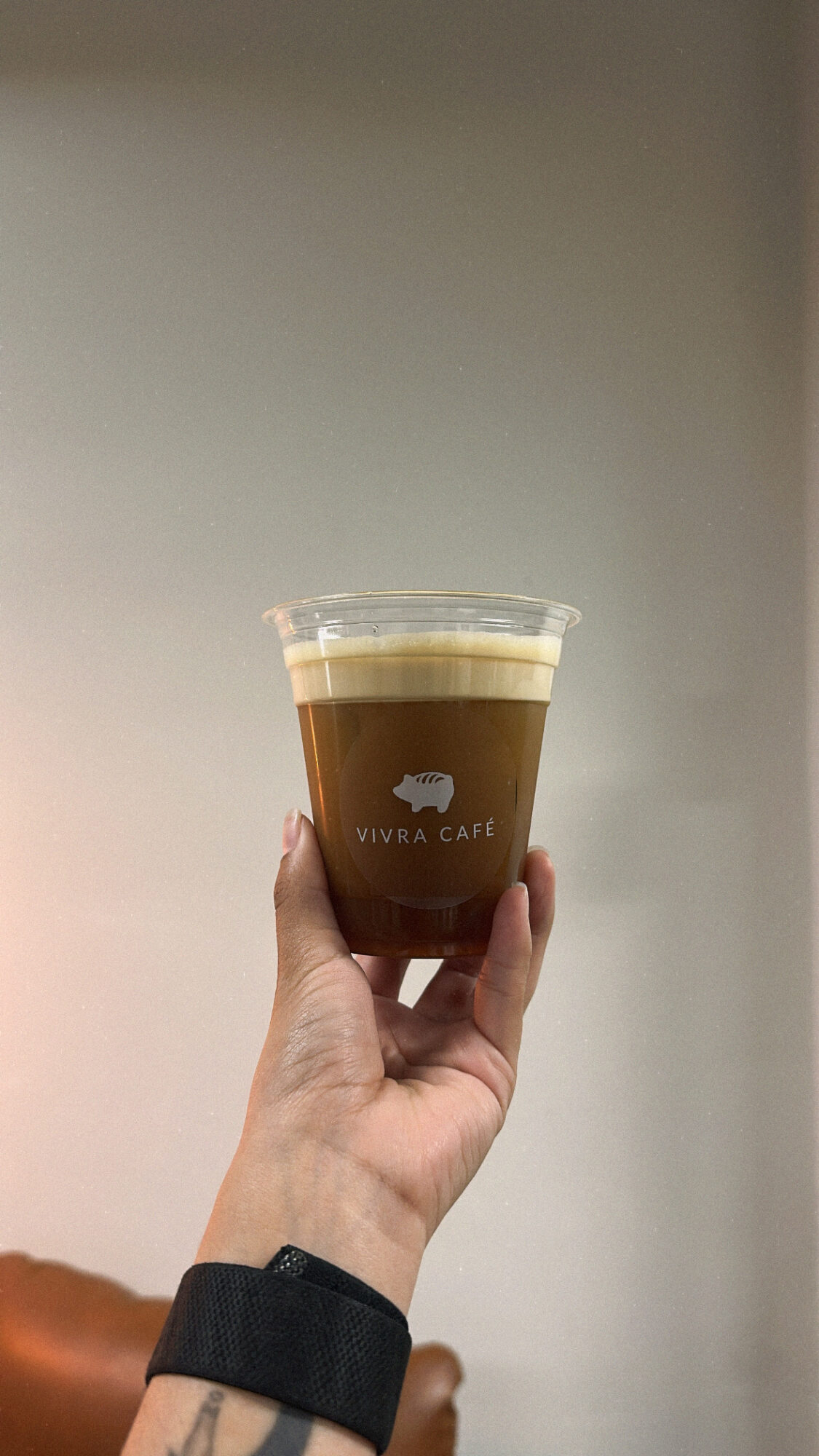 Hand holding a plastic cup of coffee with foam on top, against a plain background.