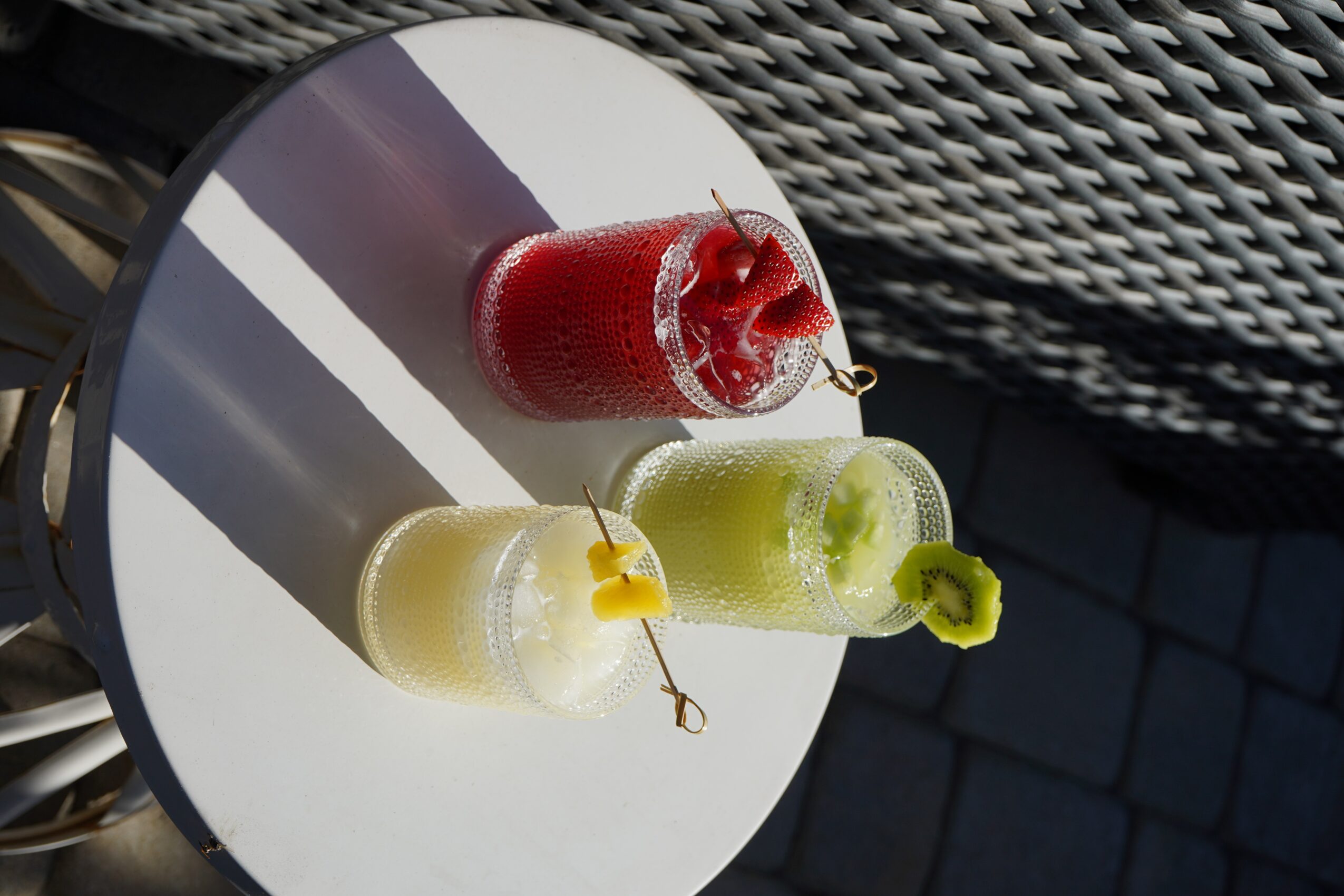 Three colorful drinks with straws on a round table, sunlight casting shadows, outdoor setting with a woven surface nearby.
