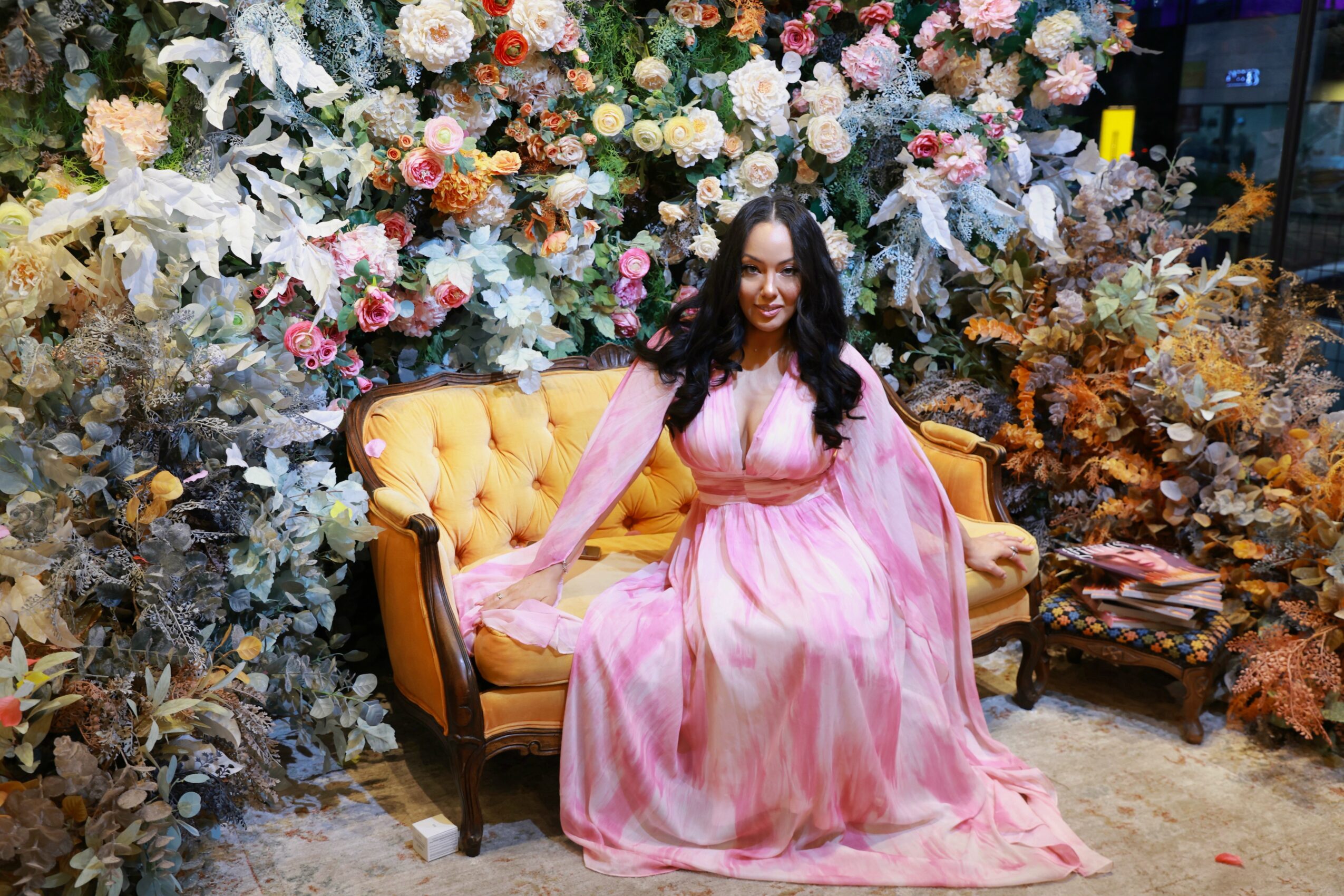 Woman in pink dress sitting on a yellow sofa in front of a floral backdrop, smiling, with a small table nearby.