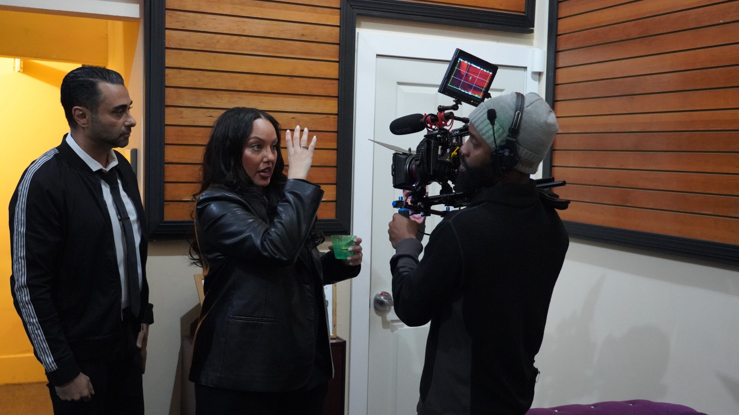Three people indoors, one woman speaking to a camera operator, two men listening, wooden wall background.