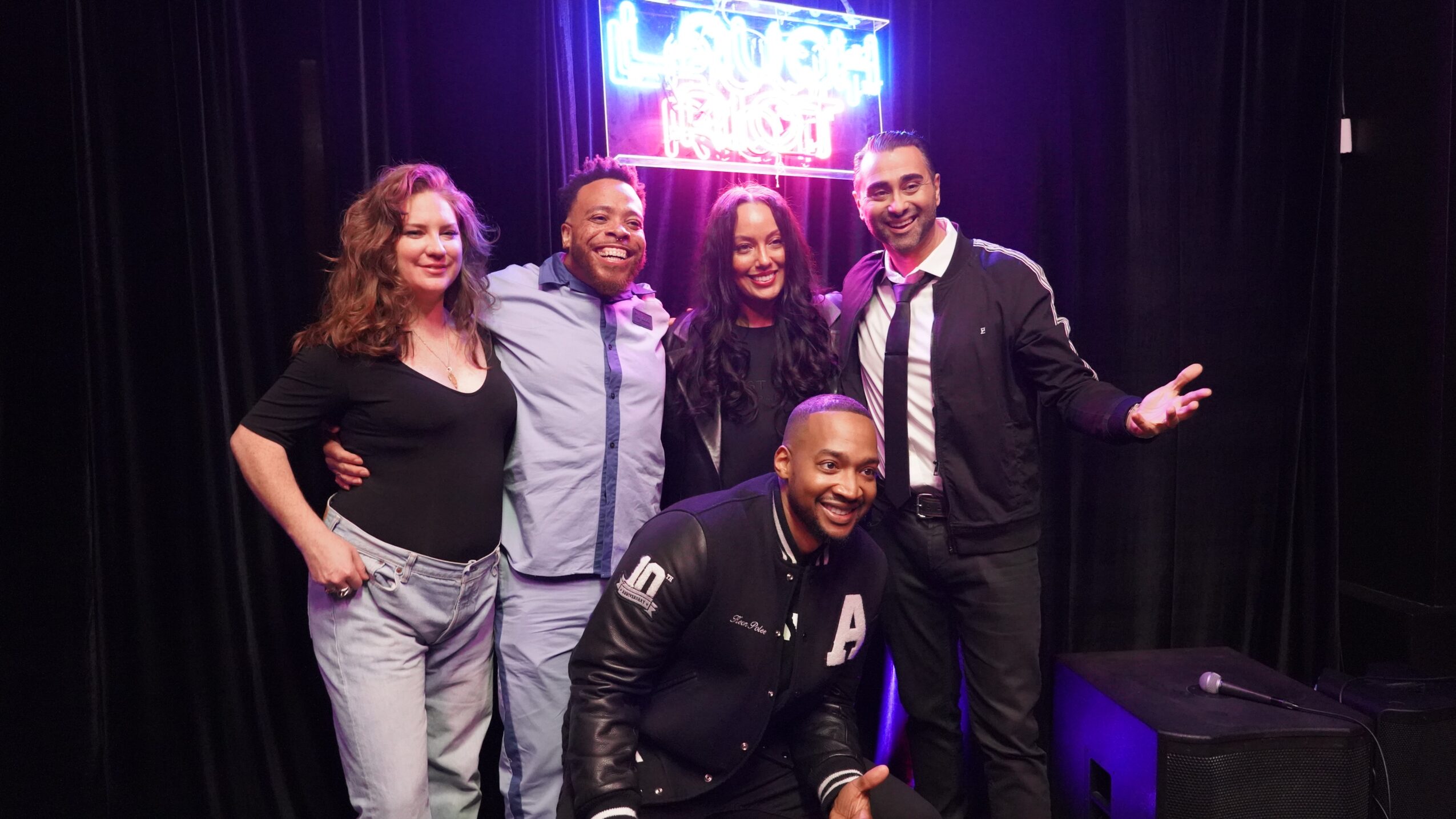 Six people posing together on stage with a neon sign in the background, smiling and dressed casually and semi-formally.