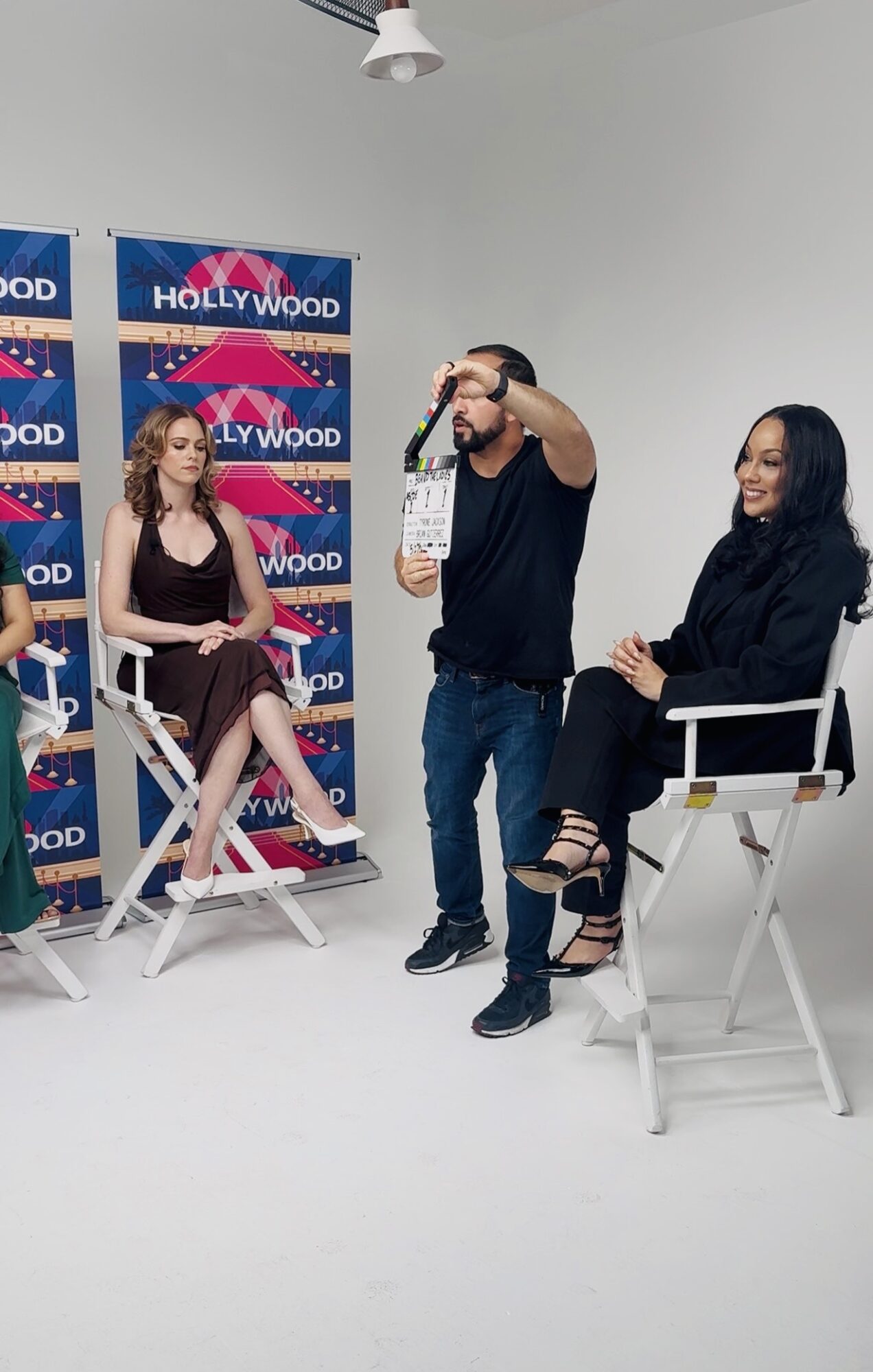 Two women and a man in a studio with Hollywood backdrop, one woman seated, the other woman and man standing, talking and laughing.