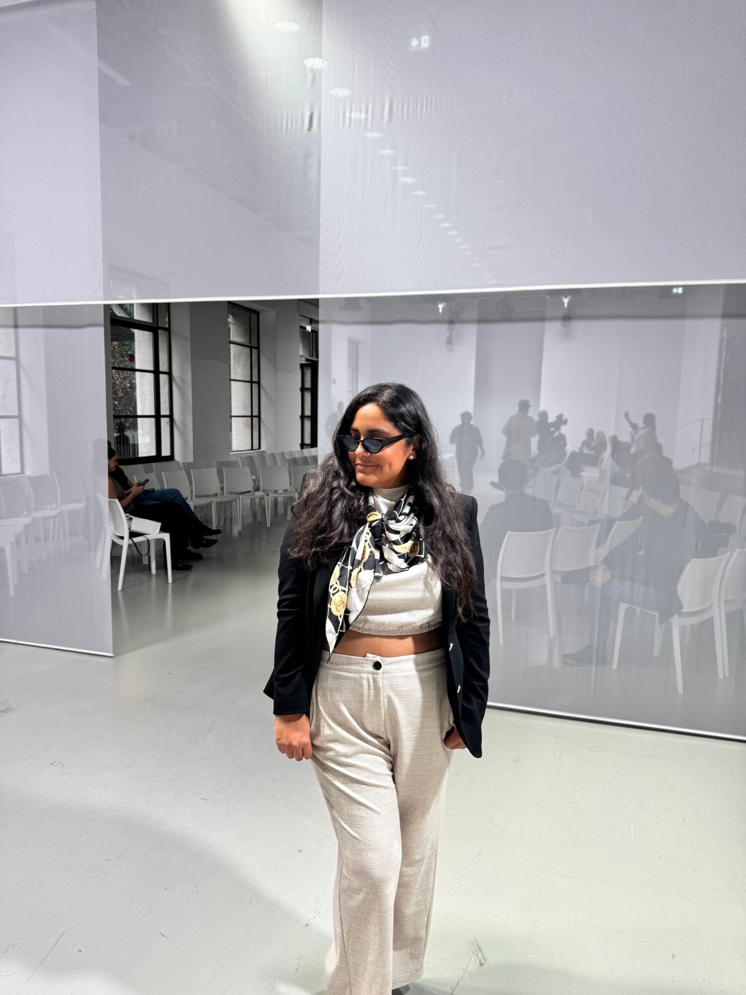 Woman standing in a modern indoor space with glass walls and reflections, wearing sunglasses, a black jacket, and light-colored pants.