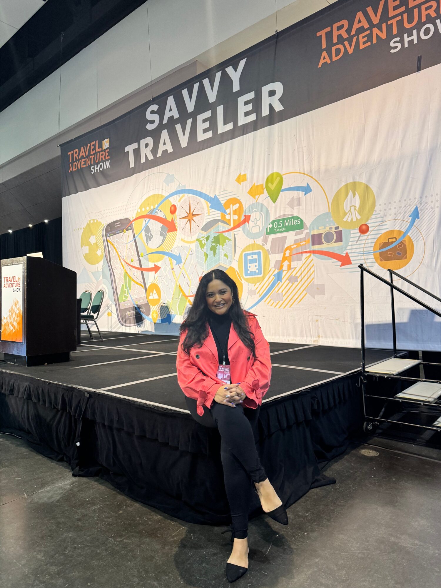 Woman in pink jacket sitting on black table at travel event with colorful travel-themed backdrop.