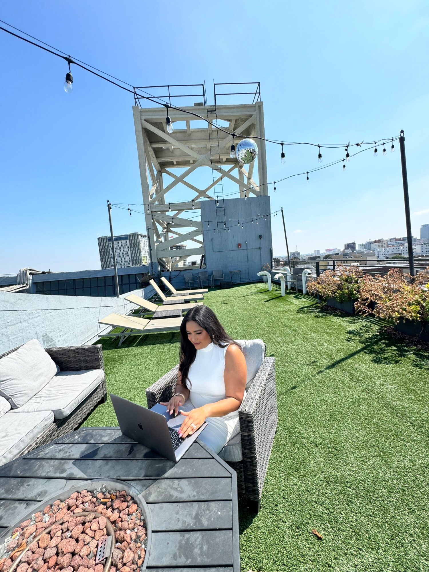 Woman sitting on outdoor bench using a laptop, with a lighthouse and blue sky in background.