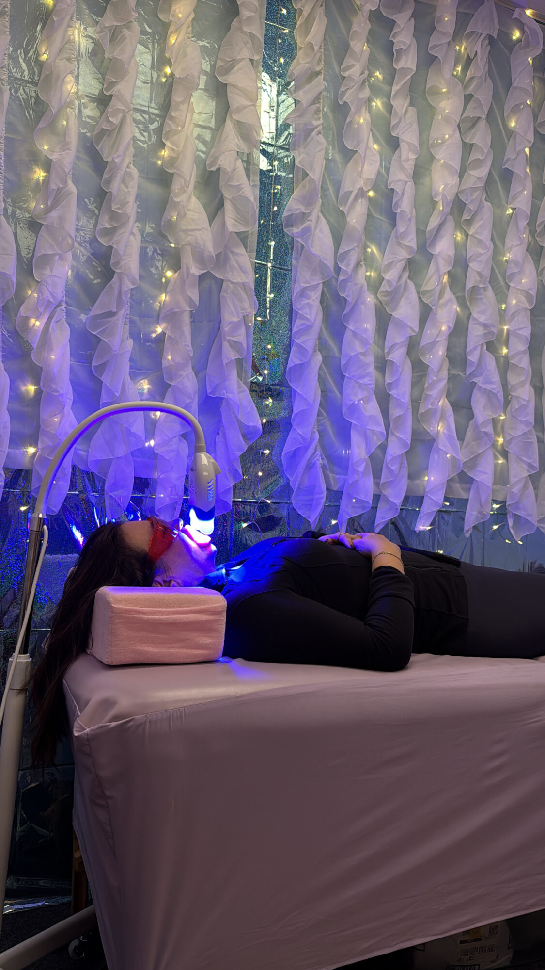 Person lying on a bed with a headrest, surrounded by decorative curtains and string lights, in a dimly lit room.