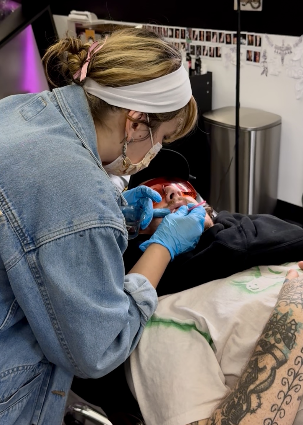 A person wearing a white headband, glasses, and blue gloves is performing dental work on a patient lying in a dental chair.