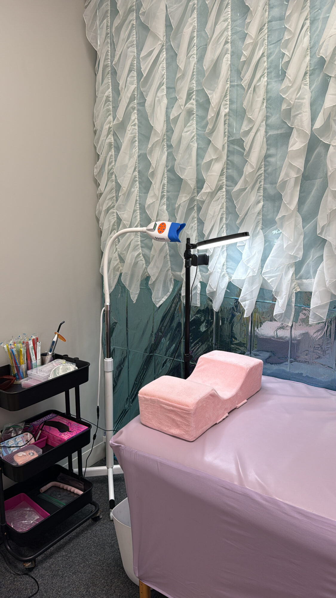 Medical examination room with a bed, pink pillow, and curtains, with medical equipment nearby.