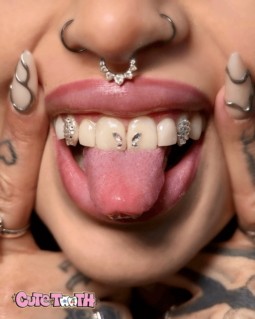 Person with tongue out, showing pierced nose and multiple decorated teeth, with tattoos on fingers and hands.