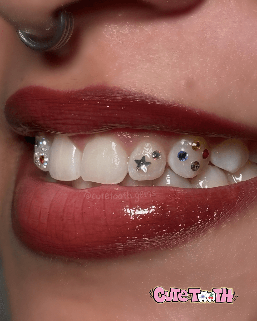 Close-up of a person's mouth with red lipstick, showing teeth with decorative star and gem stickers, and a nose piercing.