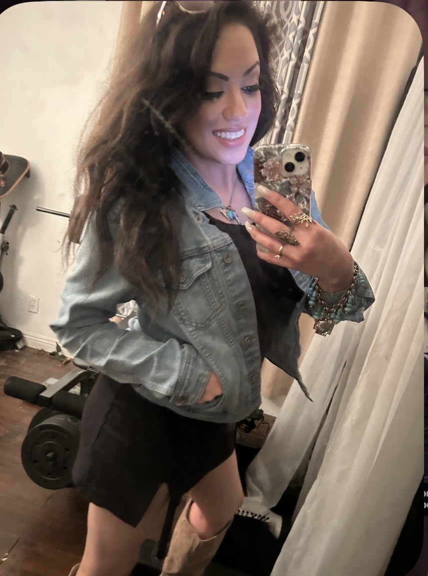 Young woman with long wavy hair smiling, taking a mirror selfie in a room, wearing a denim jacket and black outfit.