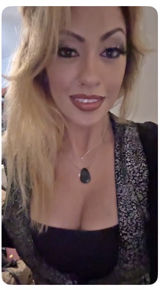 Woman with blonde hair, makeup, and a necklace, smiling at the camera, wearing a black top and patterned jacket.