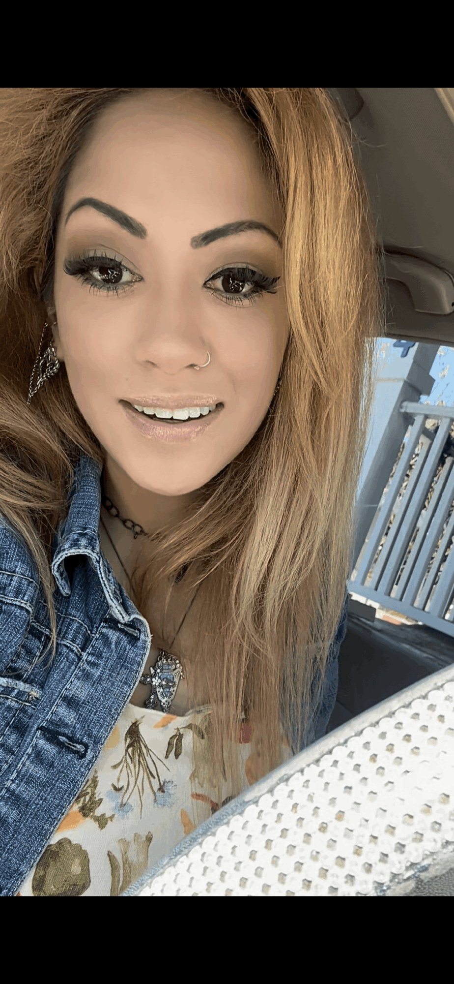 Young woman with long hair, smiling, wearing a denim jacket and floral top, sitting in a vehicle.