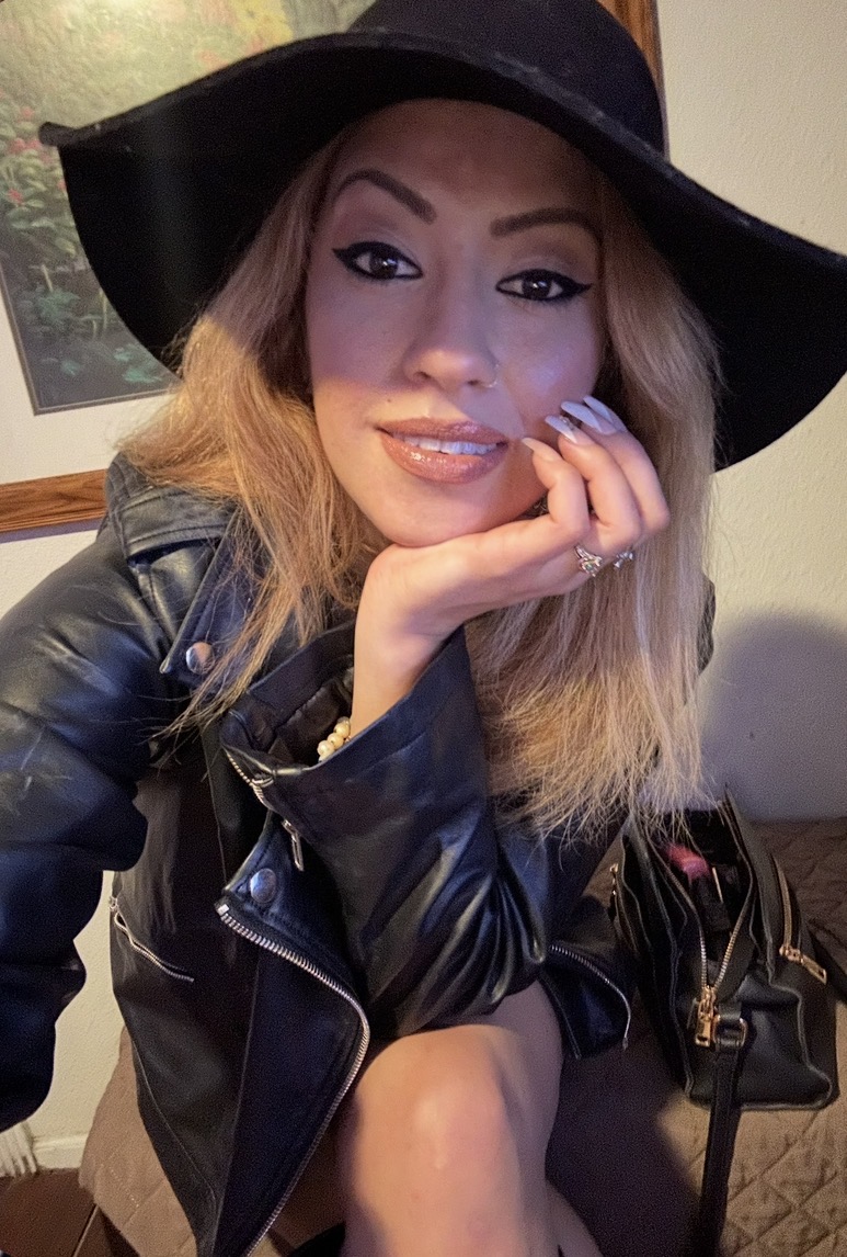 Woman with long hair wearing a wide-brimmed hat and leather jacket, smiling and resting her chin on her hand.