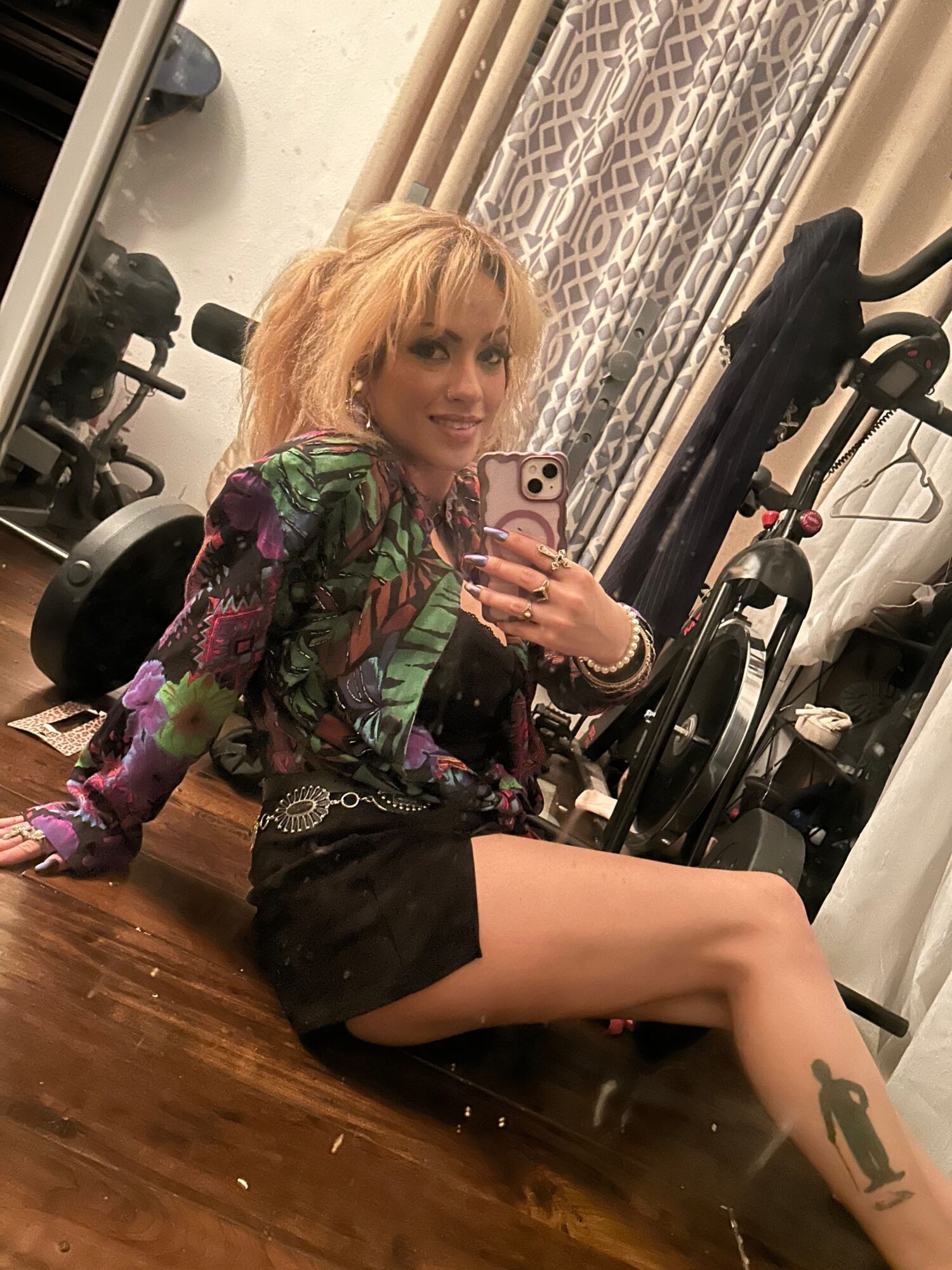 Person with blonde hair taking a mirror selfie, sitting on the floor with legs extended, in a room with curtains and equipment.