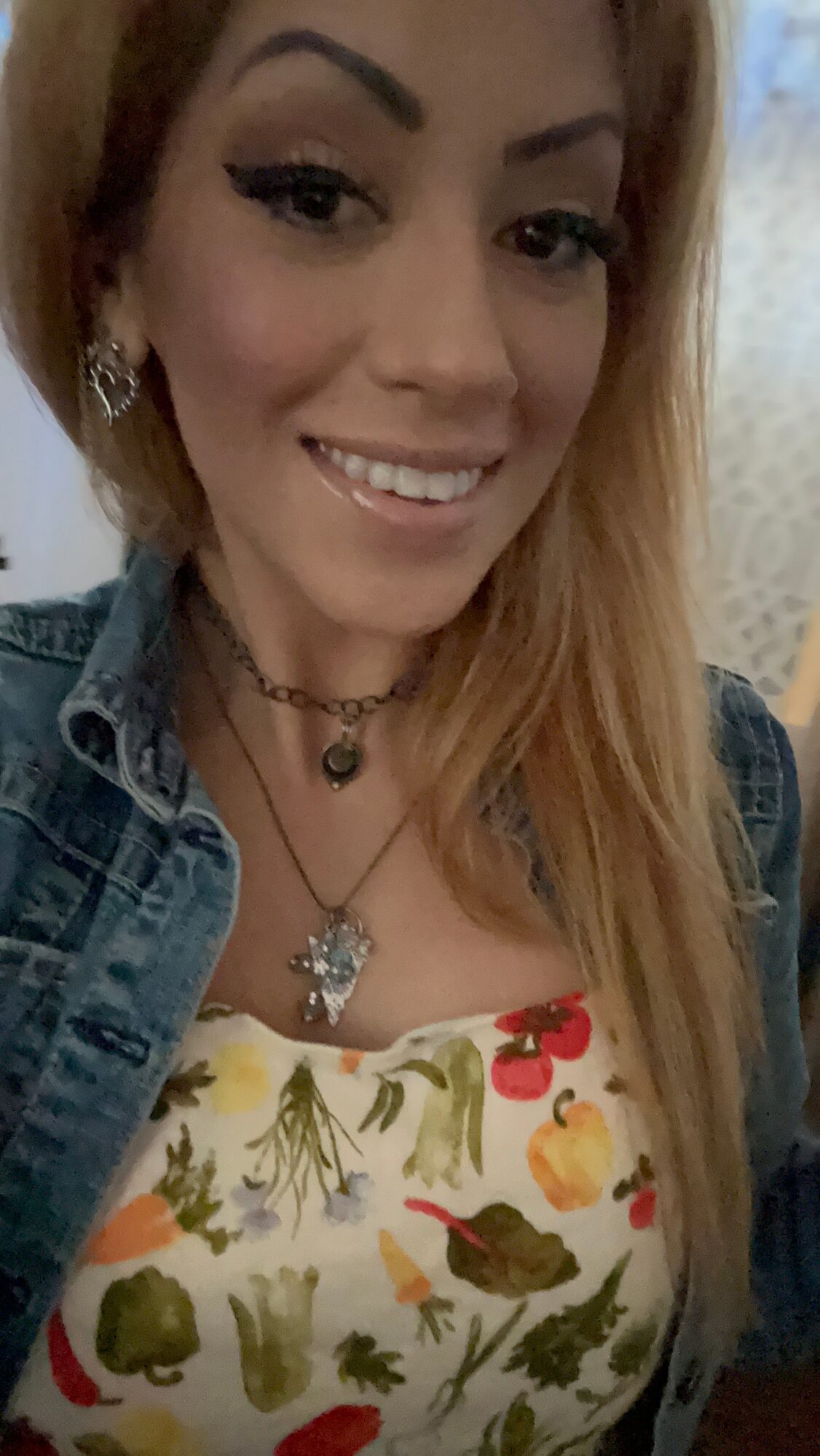 Young woman with long hair smiling, wearing a denim jacket and floral top, with jewelry and makeup.