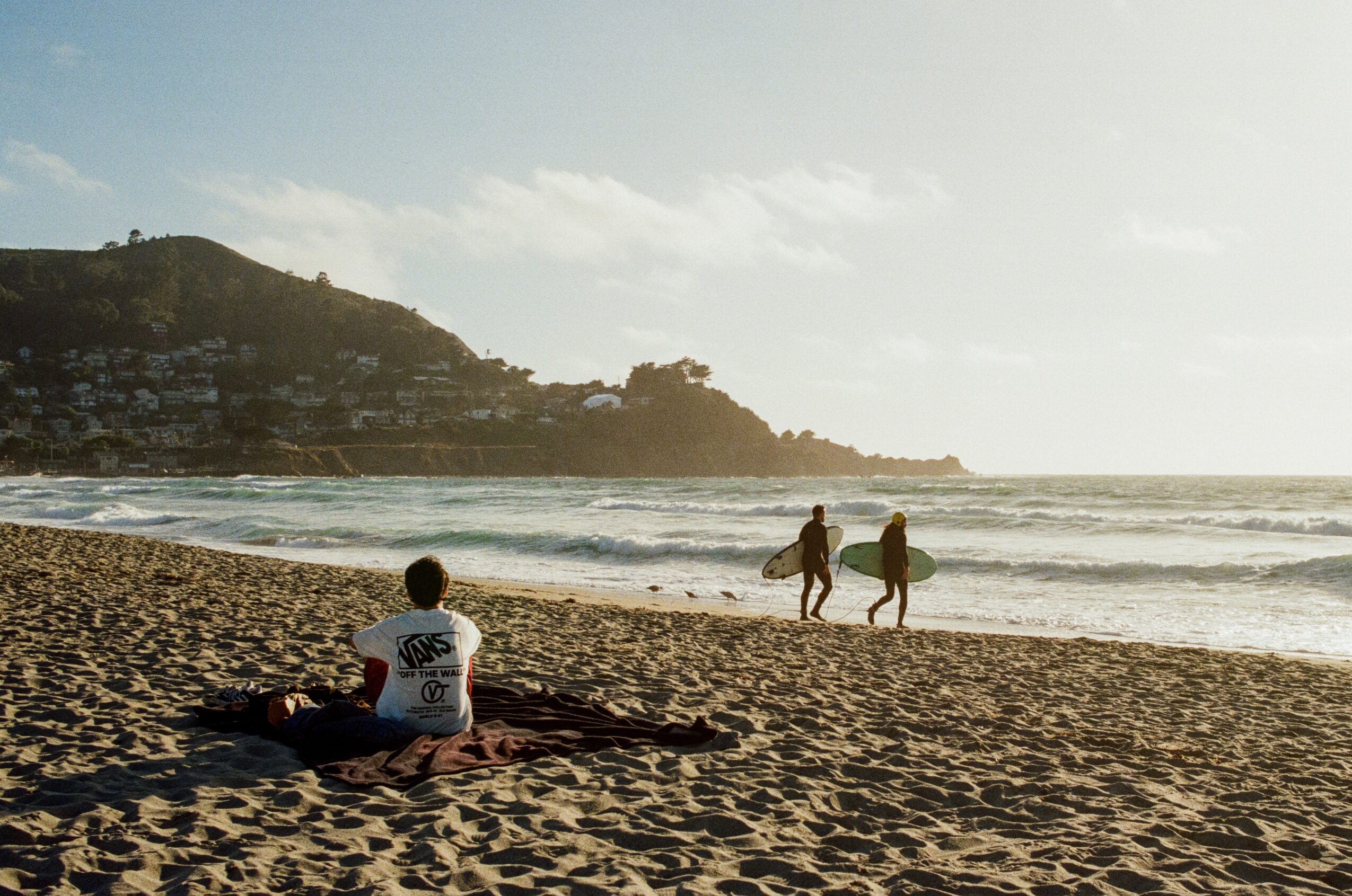 Person sitting on sand facing the ocean, two people walking along the shoreline, hill in background, sunny day.