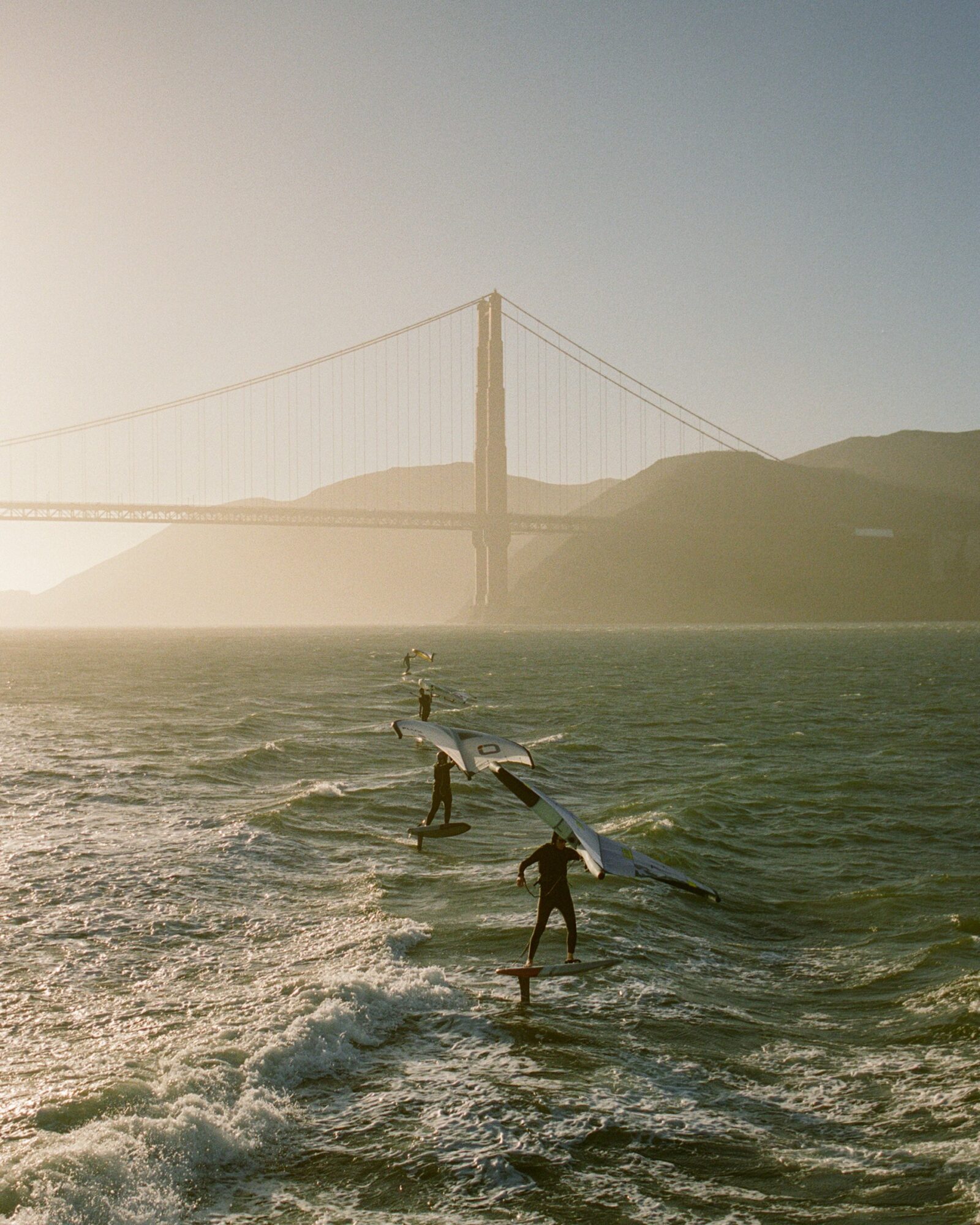 People surfing on water with a bridge and hills in the background during sunset.