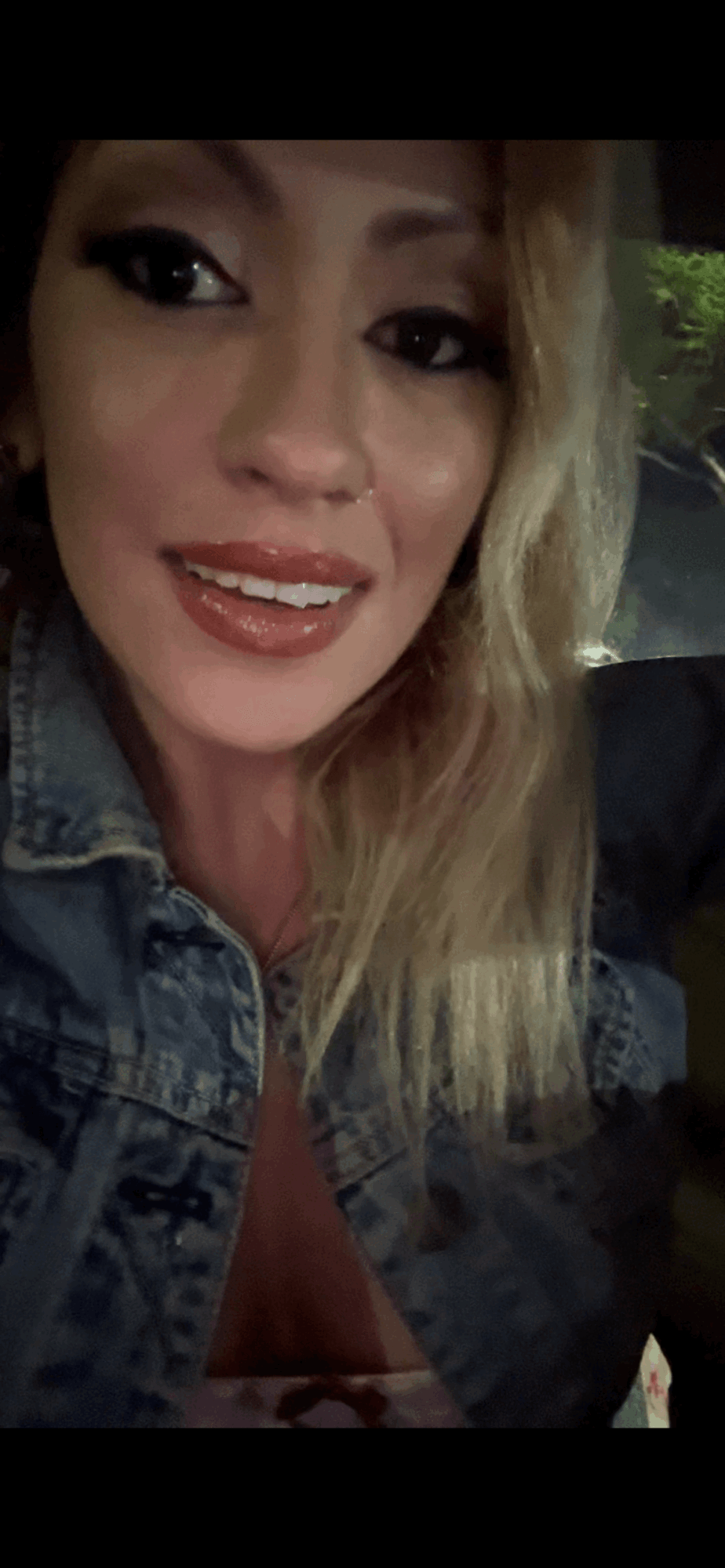 Close-up of a woman with blonde hair, smiling, wearing makeup and a denim jacket.