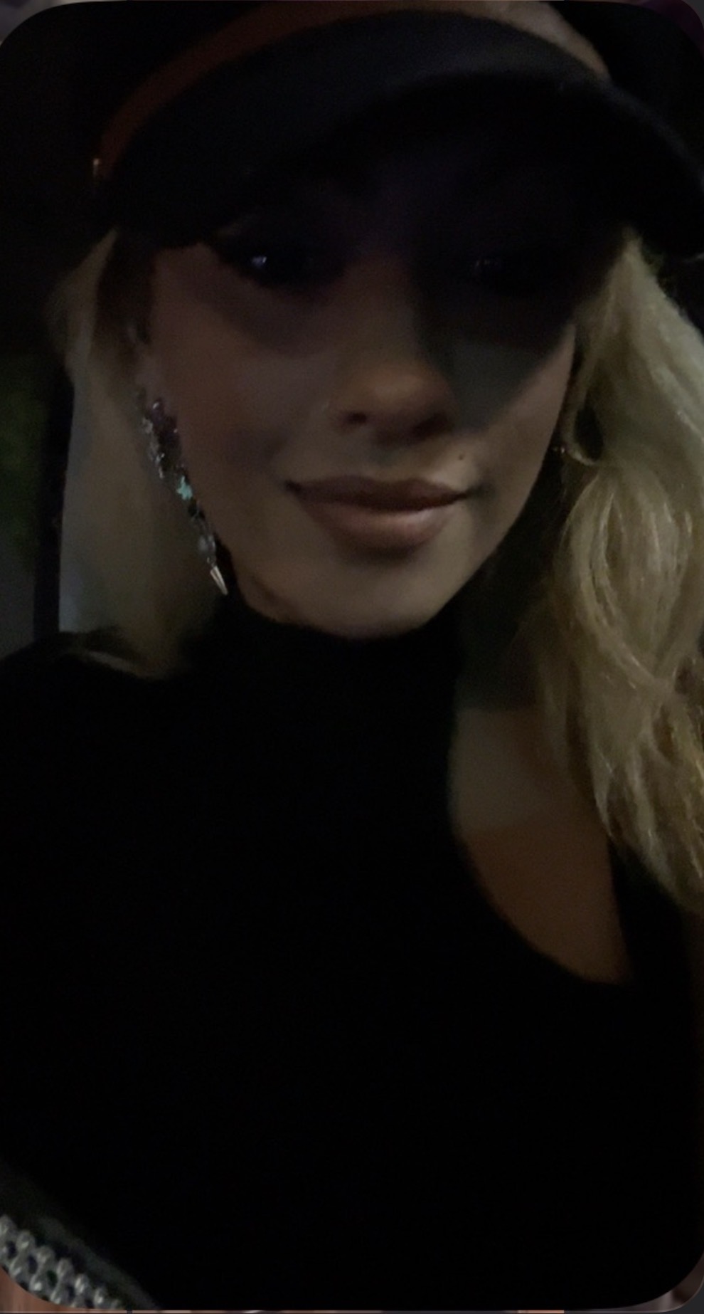 Person wearing a hat and earrings, smiling, with long blonde hair, in a dark setting.