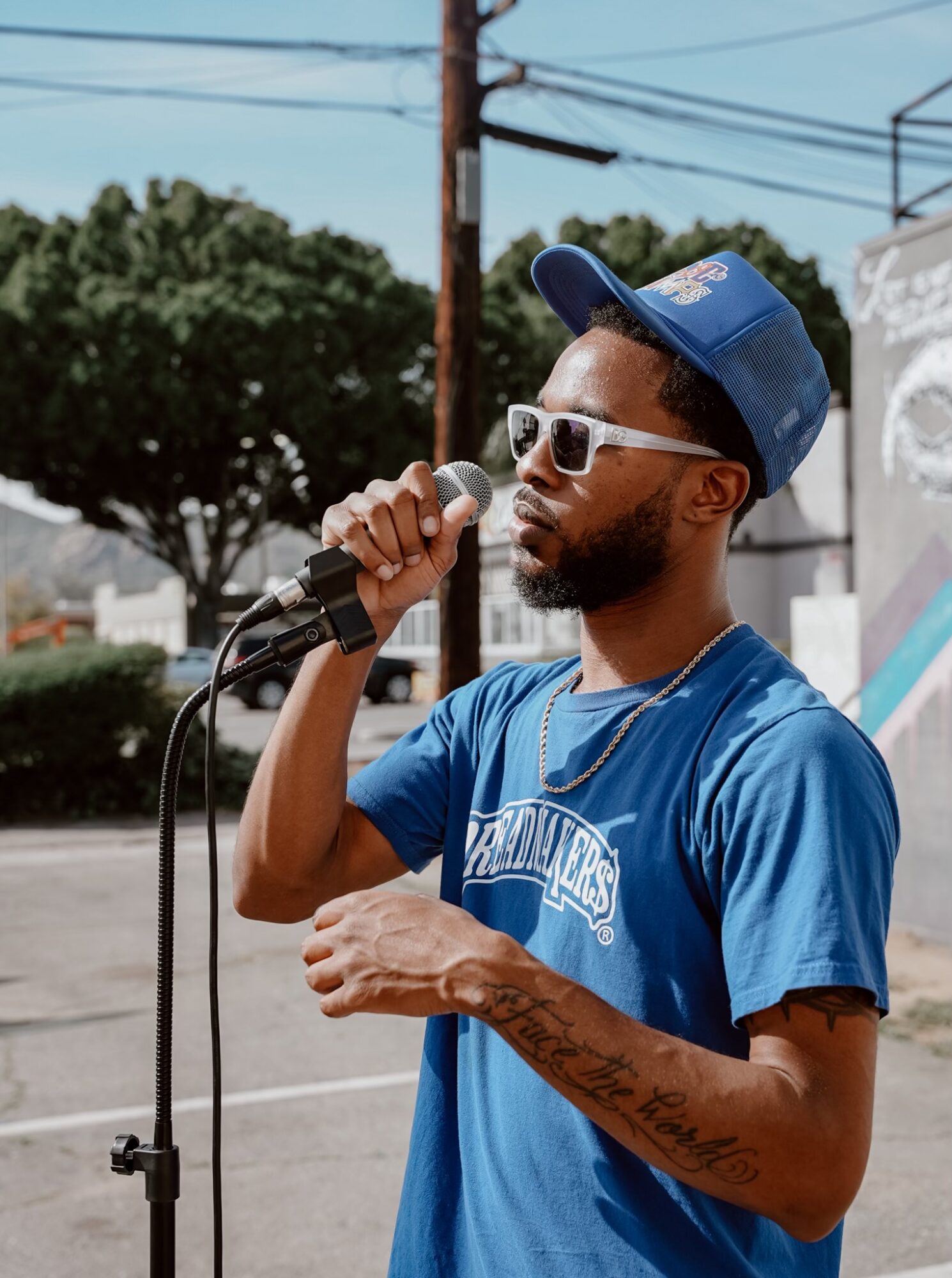 Young man with sunglasses and a blue cap singing into a microphone outdoors, wearing a blue T-shirt, with trees and power lines in background.
