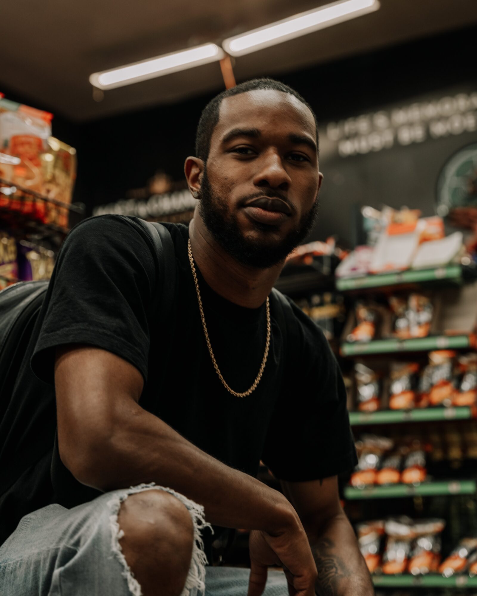 Young man with a beard and short hair, wearing a black t-shirt and gold chain, sitting in a store aisle.