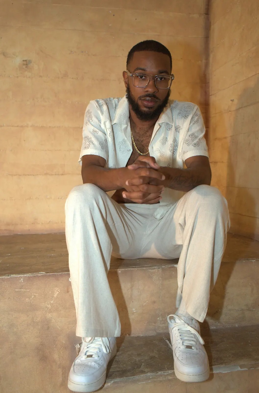 Young man sitting on stairs with hands clasped, wearing glasses, a light-colored shirt, and sneakers, in a neutral setting.