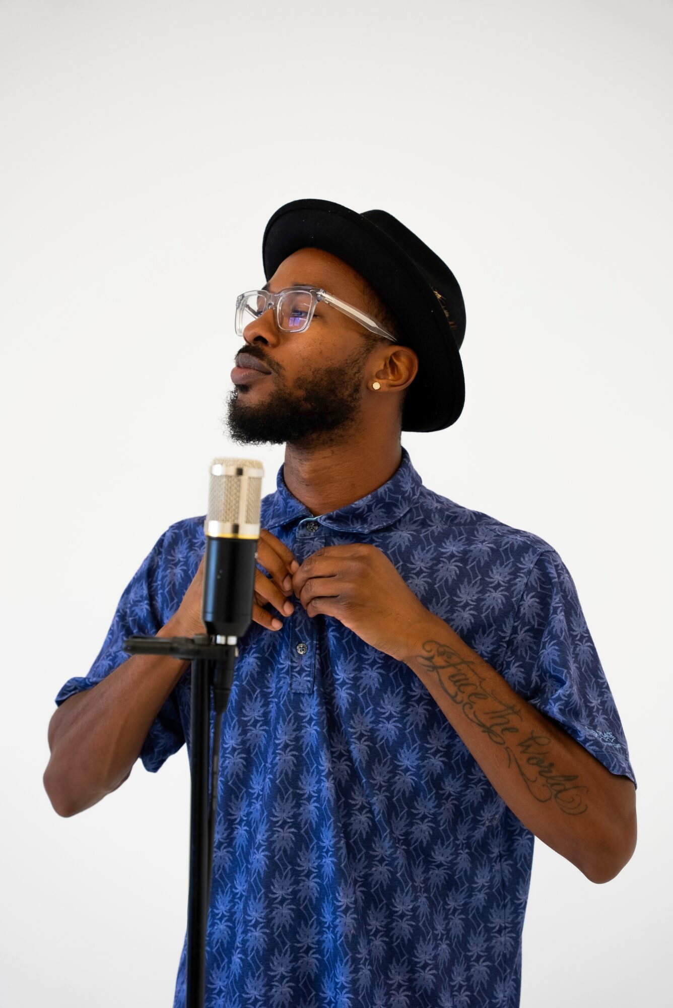 Man with glasses, hat, and beard adjusting shirt collar in front of microphone, against plain white background.